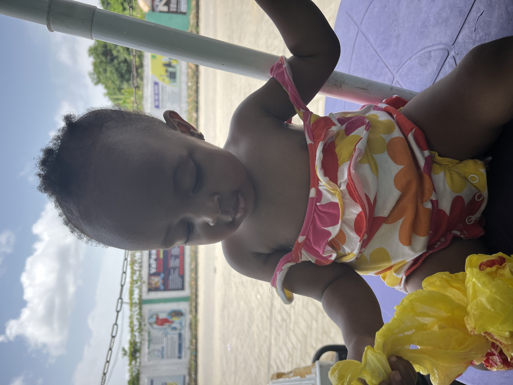 A close-up, angled, and slightly tilted shot depicts a young child with dark skin and short, curly hair, sitting outside, possibly in a public area in Lekki, Nigeria. The child is wearing a colorful floral-patterned outfit. The child is holding a crumpled yellow plastic bag in their hands.
The background includes a light-colored, possibly concrete, paved area. In the distance, signs or boards with writing and images can be seen. Above and to the left of the child, there's a white pole or beam. The sky is visible in the upper left corner, showing a partly cloudy day. A section of a purple or violet surface is also visible, likely the table top the child is seated at. The image has bright, natural lighting, casting shadows across the scene.