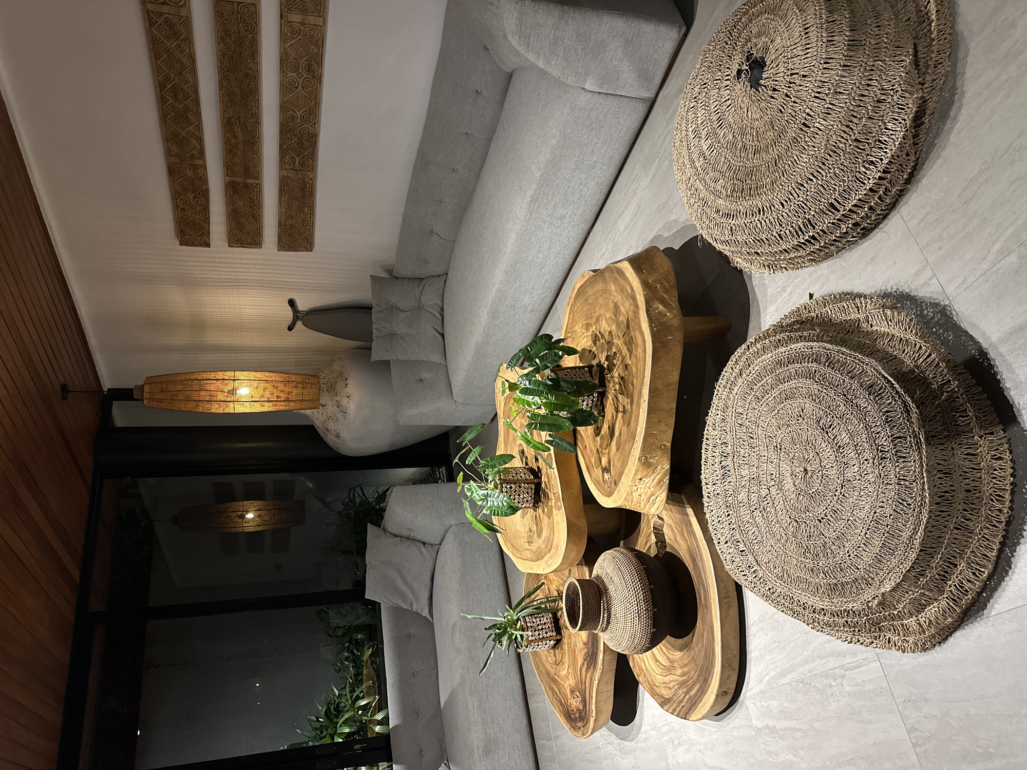 This indoor scene depicts a living room in Sukawati, Indonesia. There is a gray sectional sofa, four wooden coffee tables of varying sizes with plants on top, and two large woven straw mats on the floor. Three carved wooden panels hang on the wall. A woven lamp fixture is also visible. Light streams in from a large window.