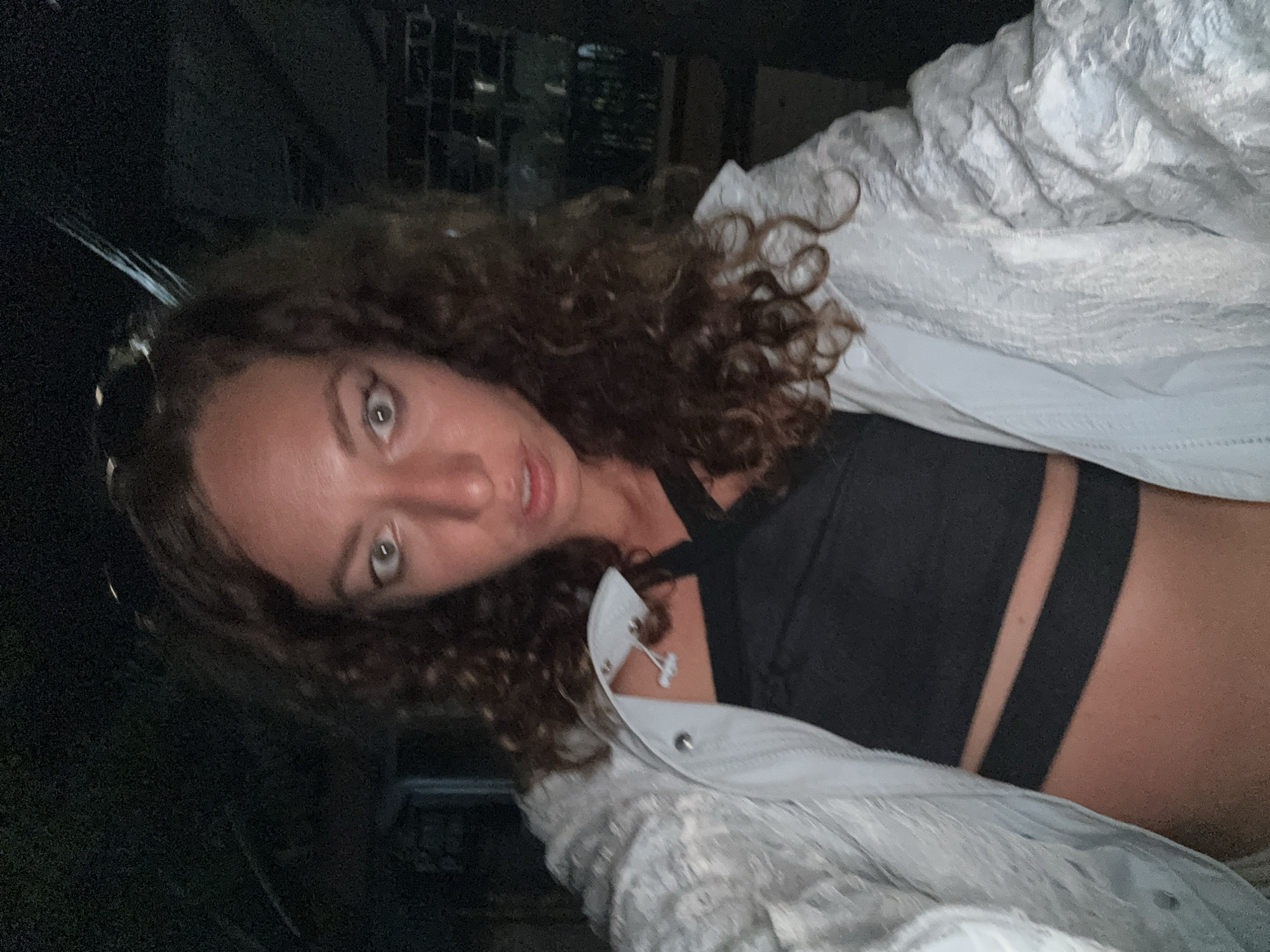 A selfie of a woman with curly brown hair and light-colored eyes is captured in a dimly lit environment, possibly at night. She is wearing a black, strappy top and a light-colored, textured jacket over it. The background is dark and out of focus, with hints of structures that could be urban buildings or outdoor elements. There are no overt signs of activity or interaction with others, suggesting a private moment or a candid shot. The woman's gaze is directed towards the camera with a curious or surprised expression. The location is not discernible from the visual information provided.