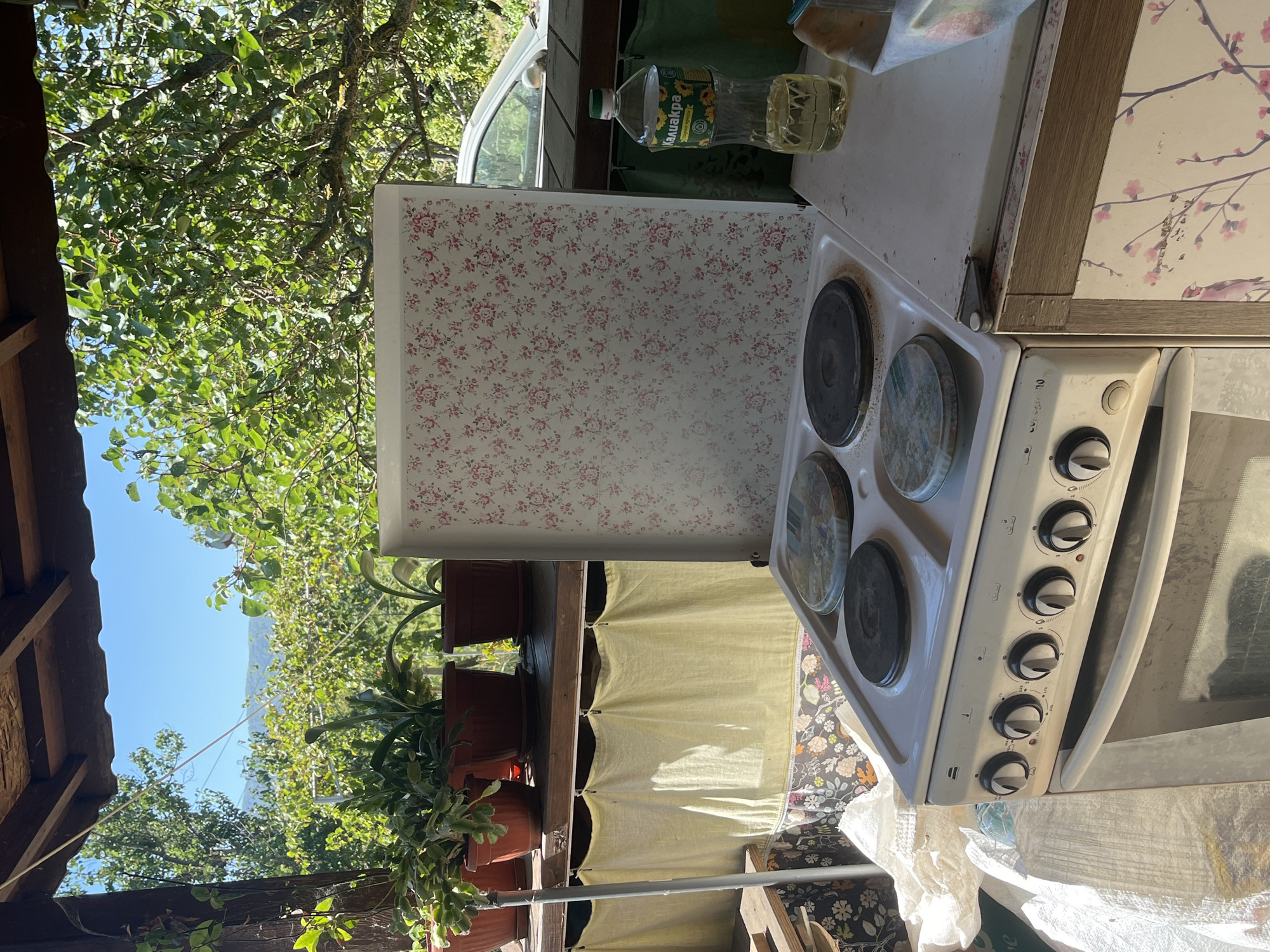 An outdoor, partially covered kitchen space in Targovishte, Bulgaria features a white electric stove with four burners and a floral-patterned lid open upwards. To the right of the stove is a clear plastic bottle filled with yellow liquid, possibly cooking oil, with a label that appears to be in Cyrillic script. Behind the stove, a car is visible through a backdrop of lush green trees and foliage under a clear blue sky. The scene is set on what appears to be a porch or veranda, with wooden beams visible overhead and several potted plants on a shelf to the left. Yellow fabric hangs as a curtain below the shelf. The overall impression is of a rustic, functional outdoor cooking area with a domestic, lived-in feel.