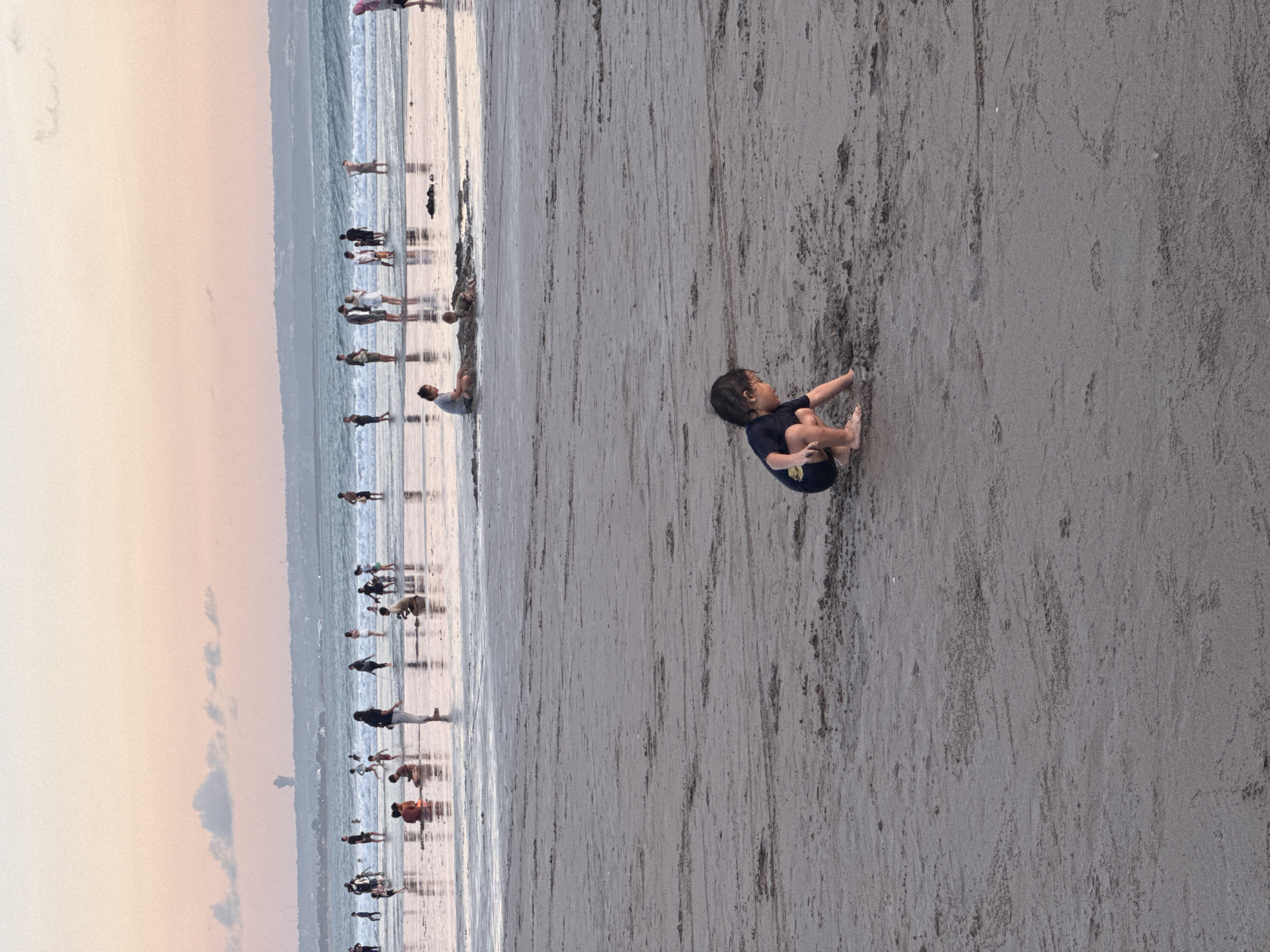 The image shows a beach at Kuta, Indonesia. A small child is playing in the sand in the foreground. In the background, numerous individuals can be seen walking and standing near the shoreline, likely along the water's edge. The sky appears to be in the early morning or late afternoon with the sun low on the horizon. The sand is damp, and the water is receding, creating a reflective surface.