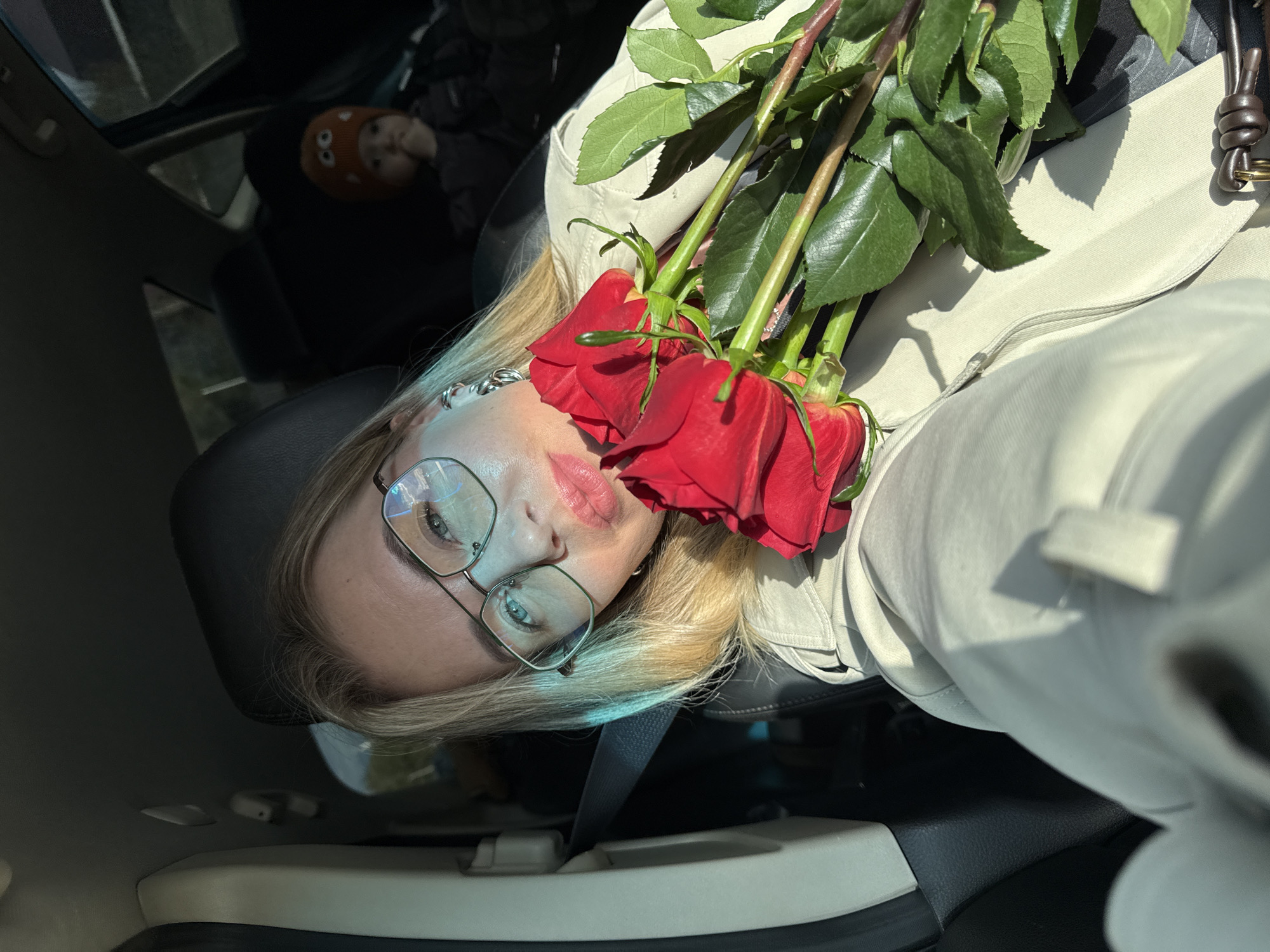 The image shows a woman taking a selfie inside a car during daytime. She has blonde hair, is wearing glasses and lipstick, and holds a bouquet of vibrant red roses with green stems and leaves to her face, partially obscuring her mouth. She is dressed in a light-colored jacket or coat, with a dark seatbelt visible across her chest. In the background, a young child wearing an orange hat with cartoon eyes is visible in the back seat of the car. The car interior features dark and light-colored elements. Bright natural sunlight streams into the vehicle, casting shadows, indicating a sunny day. No text is visible in the image.