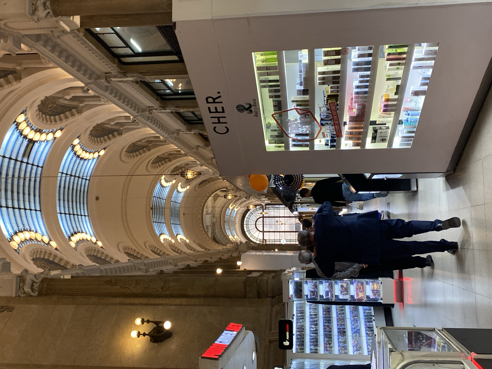The image depicts the interior of Galerías Pacífico, a shopping mall located in the city of Buenos Aires, Argentina. The view is taken from the ground floor, looking along the central corridor toward the main rotunda with its central dome. The architecture is characterized by a vaulted ceiling with detailed ornate trim and windows at the roof level. Lighting fixtures are interspersed throughout the corridor.
On the right side of the image are various commercial displays and storefronts. One prominent display is branded "CHER. Ruiz y Roca .com.ar" and showcases cosmetics and fragrances. There are several people standing near the stores, appearing to be browsing or shopping.
Other signage visible includes the brand "PUNTO GAMER" with a red neon sign. The focus is on the architectural design of the gallery and the commercial activities within. The image is oriented vertically.