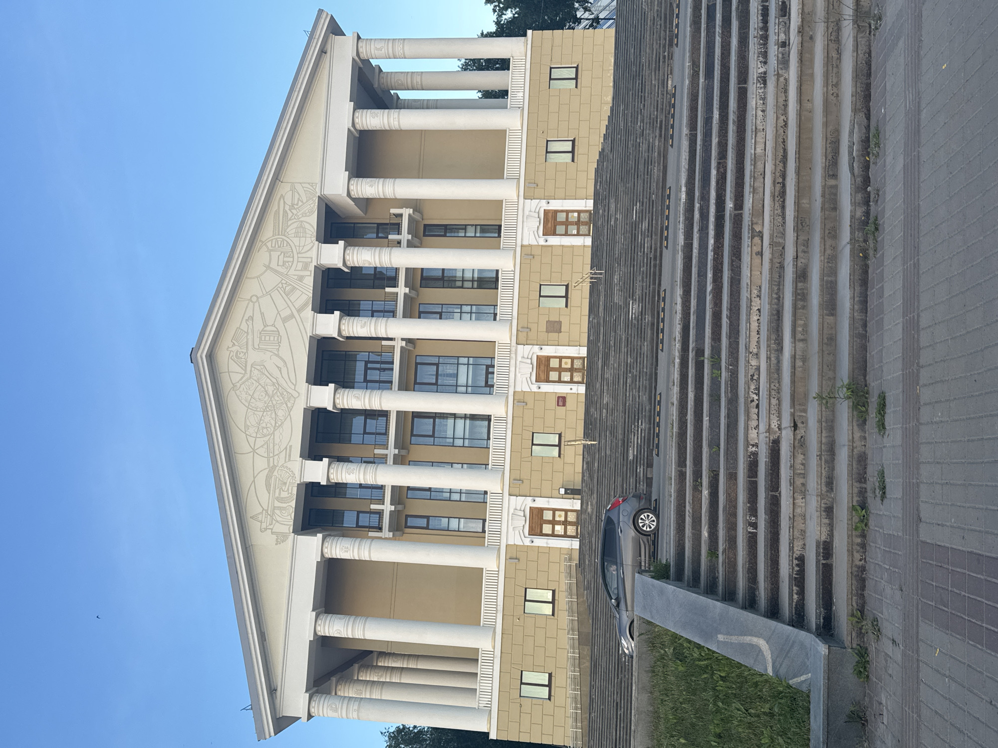 The image shows the Votkinsk State Drama Theater in Votkinsk, Russia. The theater is a large, neoclassical building with a grand facade featuring numerous white columns, a pediment adorned with relief sculptures, and a light yellow exterior. In front of the building, there is a wide, long set of stone stairs leading up to the entrance. A dark gray hatchback car is parked on a ramp to the left of the stairs, partially obscured by the steps. The sky above is clear and blue. The foreground shows a paved area with some patches of grass and weeds growing between the stones.