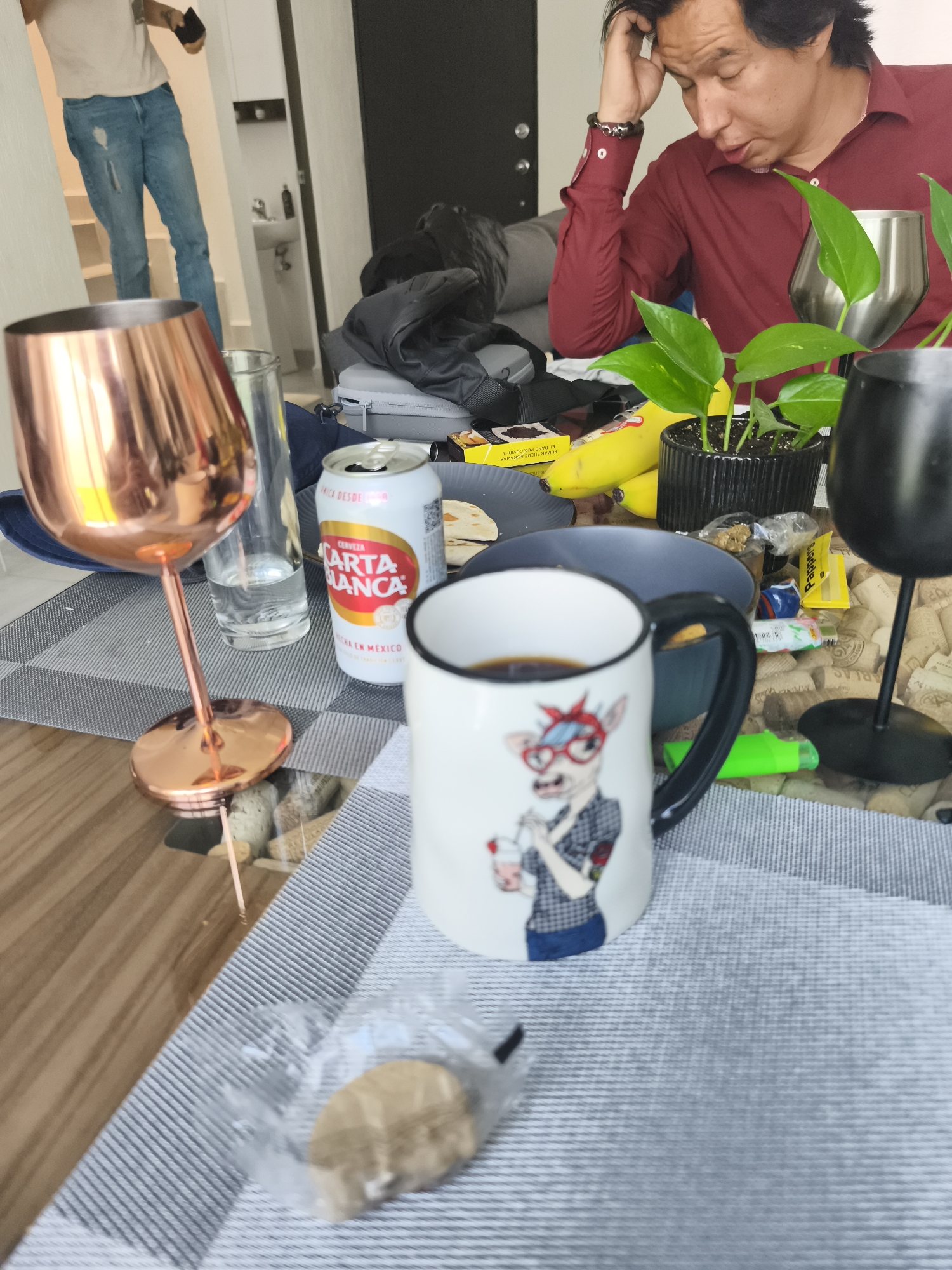 The image shows a cluttered table indoors, seemingly in a residence. A man in a red shirt sits with his head in his hand, appearing preoccupied. The table is covered with a gray and white striped placemat and holds various items including a mug with a cartoonish dog on it, a Carta Blanca beer can, bananas, a plant in a black pot, and two wine glasses, one copper-colored and the other black. Other assorted items, such as snacks and tubes, are scattered around. In the background, another man stands in a doorway wearing jeans and a light-colored shirt. The environment appears to be a modern apartment. There is no discernible evidence in the image to confirm that it is located in El Carmen, Mexico.