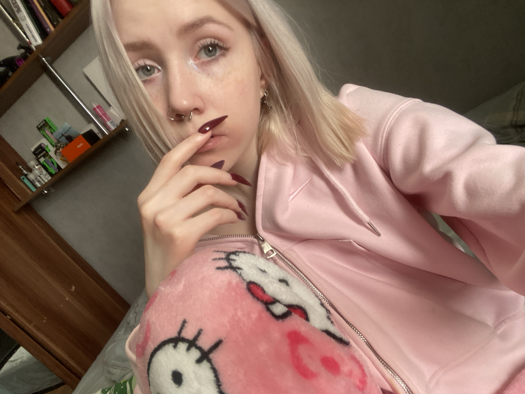 A young person, likely female, with light blonde hair, fair skin, and light-colored eyes (blue-grey) looks directly at the camera in a close-up indoor selfie. She has several freckles across her nose and cheeks. Her nose features multiple piercings, including a septum ring and a ring on her left nostril. A small metallic cross earring hangs from her right ear. She is wearing a light pink zip-up hoodie, partially open to reveal a soft, pink plush item with a white and black "Hello Kitty" like character face underneath. Her left hand, adorned with long, pointed, dark red/burgundy nails, is brought up to her mouth, with an index finger resting on her lips in a thoughtful or pensive pose.
The background on the left side of the image features a wooden bookshelf filled with books and various items, including small green and black boxes, an orange box, and clear cylindrical objects that resemble vape pens or related products. Text on one book spine appears to be "Сервер" (Server) in Russian, but other texts are indistinct. The lighting is soft and neutral, suggesting an indoor setting, likely a personal room in Izhevsk, Russia, as per the provided context. The overall impression is casual and personal.