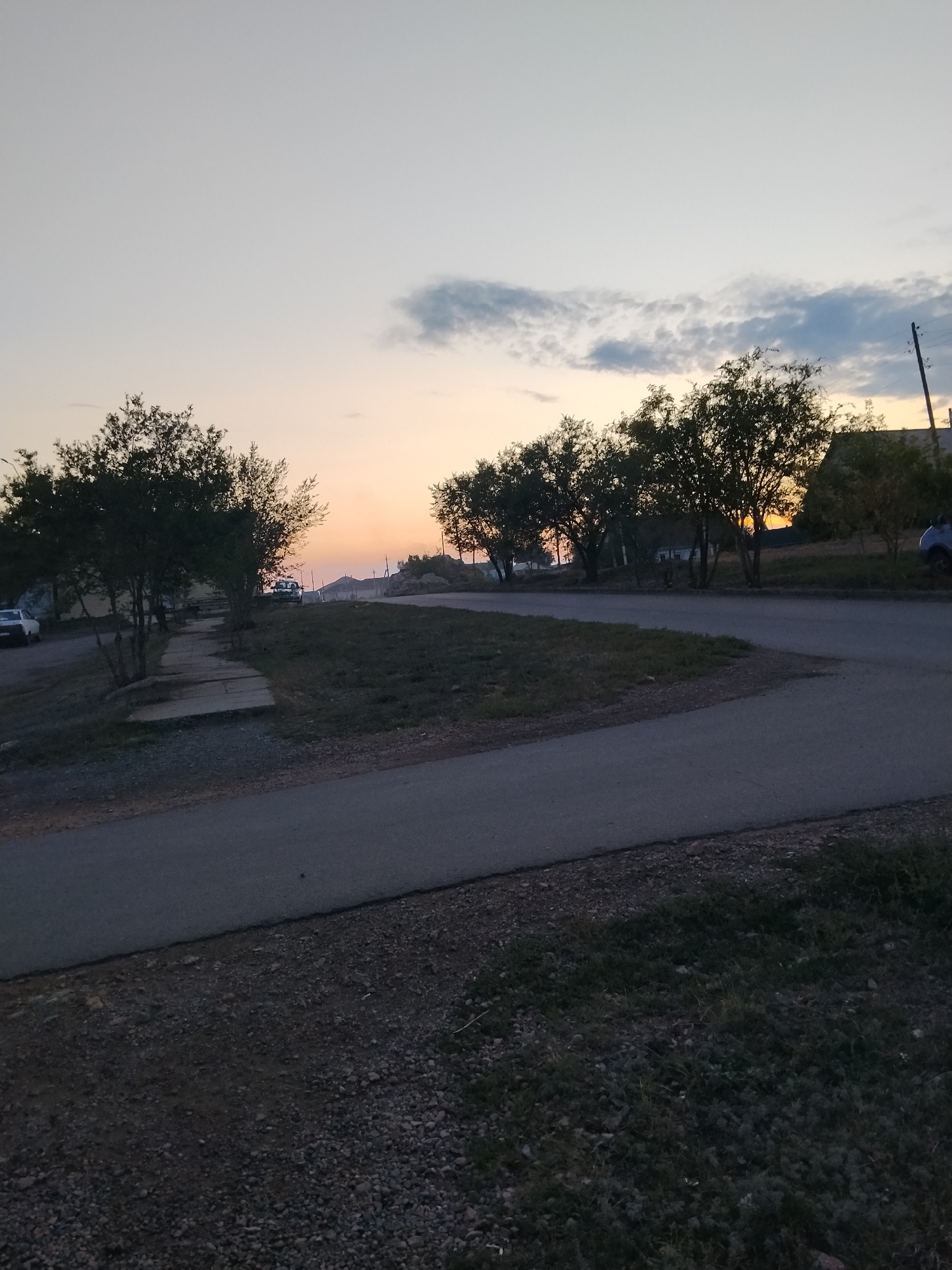 This image depicts a twilight scene on a road in Kokshetau, Kazakhstan. The sky is a gradient of light blue and pale orange, with a few scattered clouds. Several trees with sparse foliage are visible, silhouetted against the sky. A paved road curves through the scene, with a gravelly shoulder and a grassy area. On the left side of the road, a sidewalk with concrete slabs is present. Two white cars are parked on the left side of the road. In the distance, buildings and utility poles are visible. The overall impression is a quiet, rural road at dusk.
