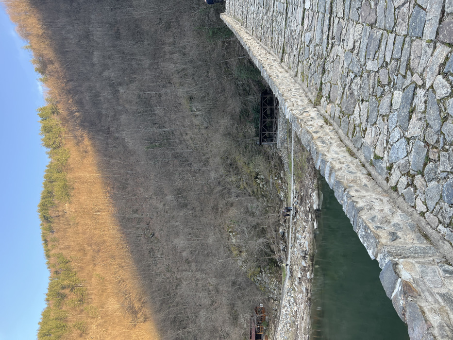 The image captures a valley scene in Diadovtsi, Bulgaria, under clear skies. A stone arch bridge, constructed from irregularly shaped grey stones, extends from the lower right to the mid-left foreground, forming a low wall along its visible side. The bridge's surface is paved with similar stone slabs. A body of dark green water is visible to the left of the bridge, bordered by a rocky and muddy bank.
In the mid-ground, along the far bank of the water, a dirt path is visible. A group of three individuals walks along this path towards the left. Further to the left on the same path, a single person is also visible. Near the left edge of the frame, a small wooden cabin with a red roof stands among sparse trees on the bank. Another larger, open-sided wooden structure with a dark roof is situated on the near bank, further into the valley from the bridge's perspective.
The background features steep hillsides covered predominantly with leafless deciduous trees, suggesting a dormant season. A distinct diagonal shadow from an unseen peak bisects the hillside, illuminating the upper portion with golden sunlight that also highlights a line of evergreen pine trees along the ridge. The lower portion of the hillside remains in shadow, revealing rocks and sparse vegetation. One individual is partially visible walking on the stone bridge towards the upper right edge of the frame.