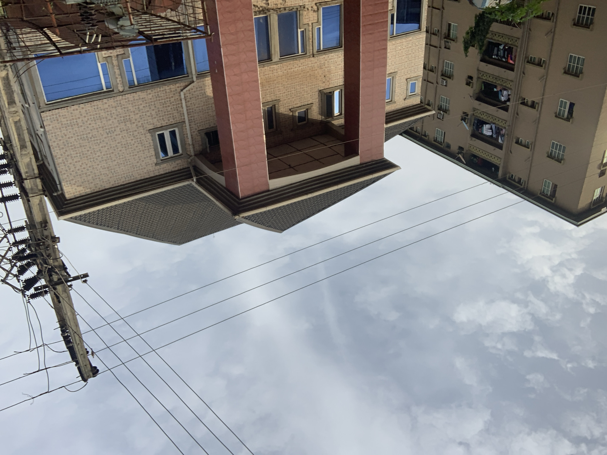 This is a mid-range shot of residential buildings and a utility pole in Nkpor, Nigeria. On the left, a tall, tan apartment building with numerous windows, balconies with clothes drying, and air conditioning units. In the center and right, a newer, multi-story building with a gabled roof and prominent red pillars. This building features brick-like siding and large blue-tinted windows. A utility pole with multiple wires and insulators stands to the right of the newer building. The sky is overcast with scattered clouds.