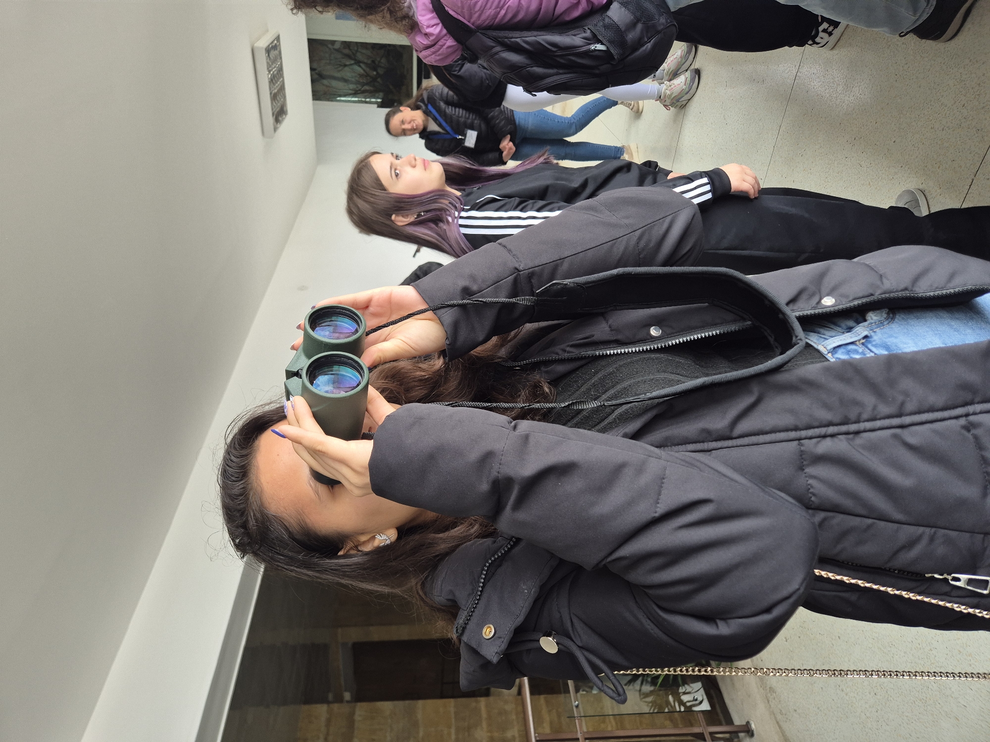 A group of people are standing indoors, likely at a visitor center or educational facility in Srebarna, Bulgaria. One person, a woman with long dark hair and wearing a black puffer jacket and blue jeans, is holding binoculars up to her eyes. Another person is standing directly behind her, wearing a black tracksuit with white stripes. Other people are also in the background. The building has white walls. A glimpse of outdoor scenery can be seen through a window behind the group.
