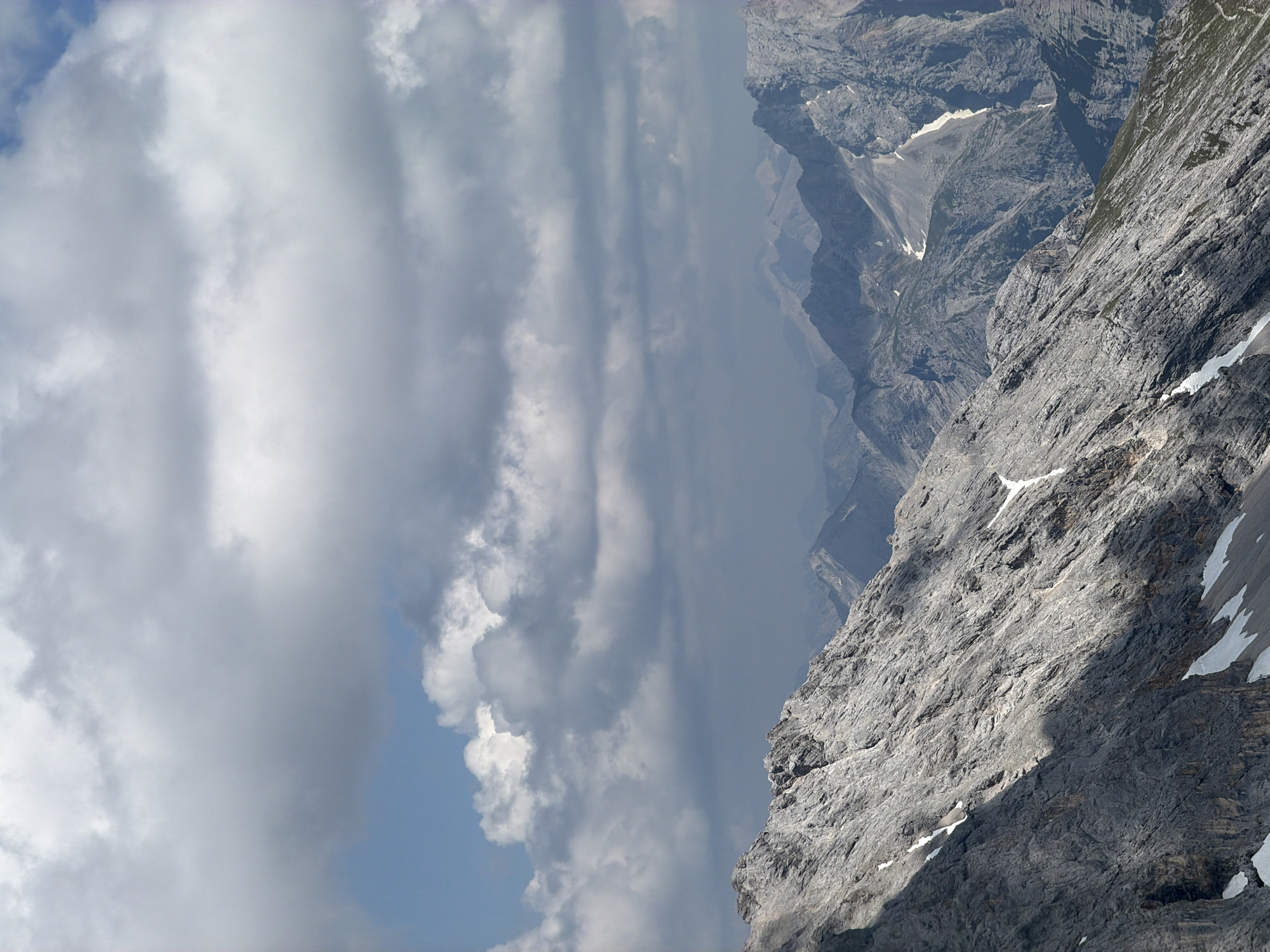 This is a vertical, wide-angle, close-up shot of a rocky mountain landscape with dramatic clouds. The foreground is dominated by a steep, grey, rocky slope, deeply textured with fissures and shadows. Patches of white snow or ice are visible on the slope, particularly near the top right. A diagonal shadow cuts across the rock face, creating a stark contrast between sunlit and shaded areas.
In the background, successive layers of rugged mountain ranges recede into the distance, bathed in a hazy, atmospheric light. These distant peaks are also rocky and show signs of geological layering.
The sky is filled with large, voluminous clouds. On the left, the clouds are bright white and fluffy, with glimpses of a clear blue sky peeking through. As the clouds move towards the center and right of the image, they become darker and more diffuse, appearing as a thick, grey veil that partially obscures the distant mountains.
The lighting suggests it is daytime, likely late morning or early afternoon, given the angle of the shadow. The weather appears to be mixed, with clear sky and bright sunshine juxtaposed with heavy cloud cover, possibly indicating an approaching storm or recent rain.
There is no visible text in the image. The overall mood is one of grandeur, ruggedness, and the power of nature, with a sense of immense scale and height. The image evokes a feeling of being high up in the mountains, perhaps on a hiking trail or a scenic overlook. The location context of Garmisch-Partenkirchen, Germany, suggests this is likely a view of the Bavarian Alps.