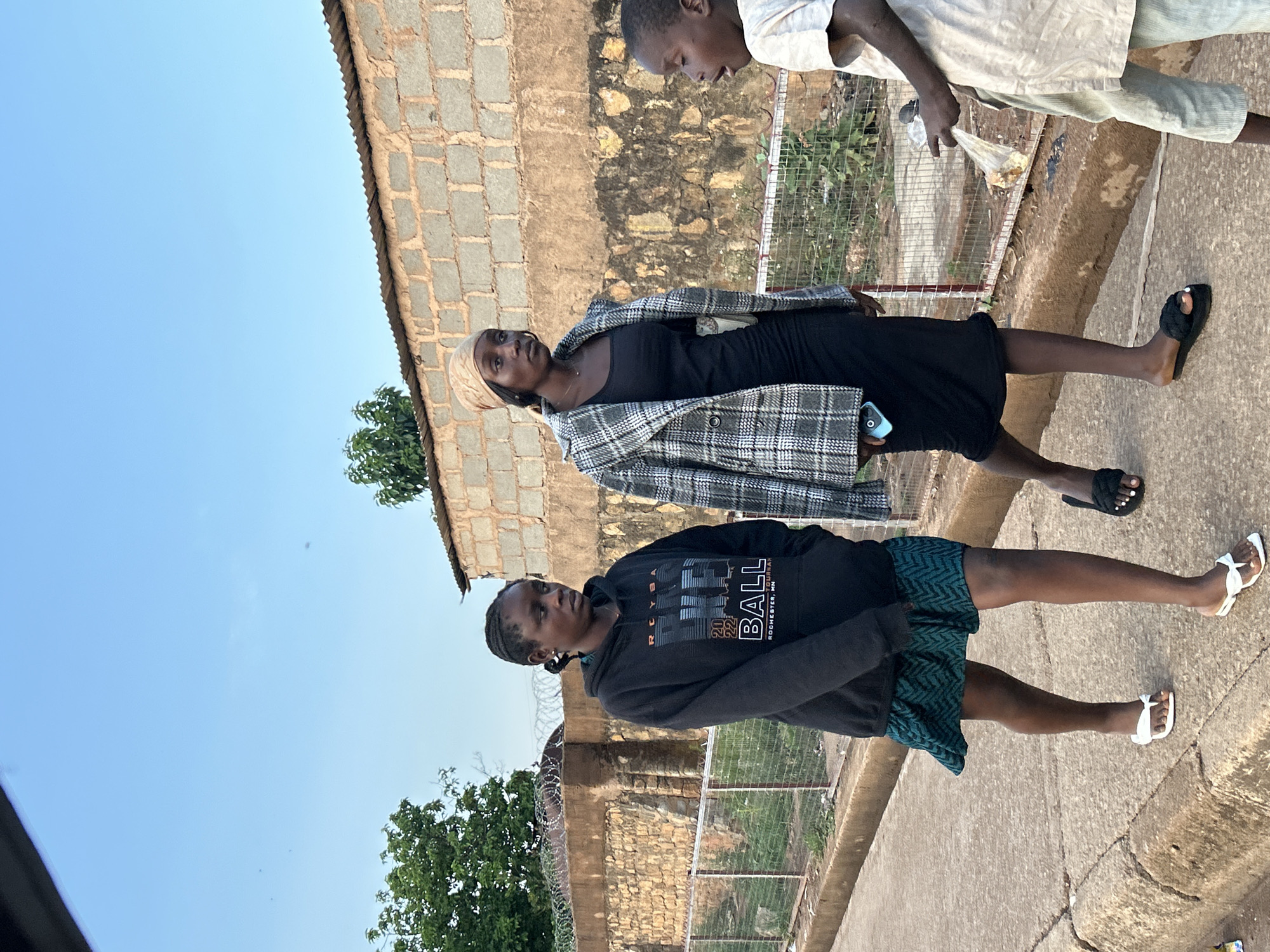 The image depicts three people on a street in Bukuru, Nigeria. Two women stand together: one wearing a black dress and a plaid jacket with a head covering, and the other wearing a dark sweatshirt and patterned shorts. A third person, a male, stands slightly behind them, partially visible. The setting appears to be a residential area with buildings made of stone and metal wire fencing. The sky is blue and there are trees visible in the background. The ground is paved with concrete.
