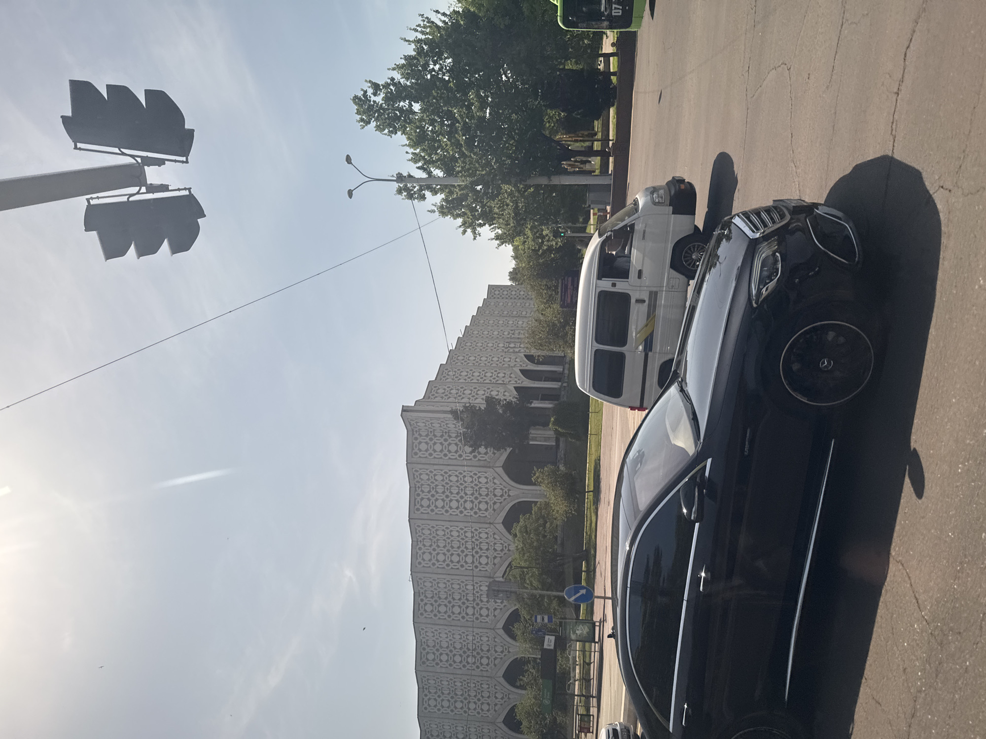 The image captures a street scene in Tashkent, Uzbekistan, showcasing a mix of urban infrastructure and vehicles. A black sedan occupies the foreground, parked on the right side of the street. Next to it, a white van is also parked. A large building, likely a government or public institution, is visible in the background; its facade is adorned with an intricate geometric pattern. The sky is bright and relatively clear, with a few scattered clouds. A traffic light structure is present in the upper left corner of the image. Street lamps are also noticeable along the street.
