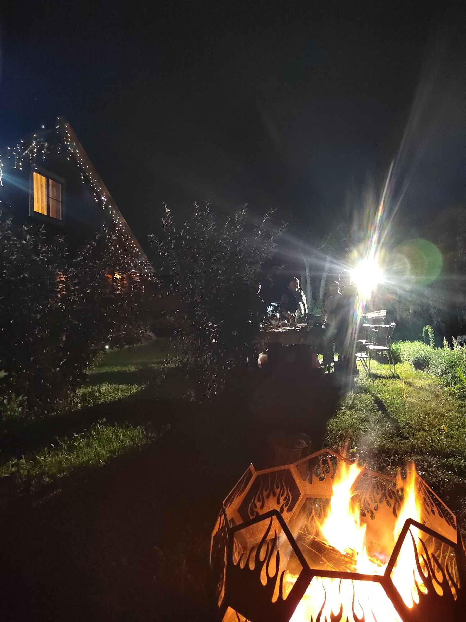 This is a night photograph taken outdoors, likely in a backyard or rural setting. In the foreground, a hexagonal fire pit with flame cutouts burns brightly, casting a warm glow. To the left, a wooden house with a steep roof is decorated with strings of small, white lights. One window of the house is illuminated. In the mid-ground, a group of people are gathered around a table, partially obscured by trees and foliage. A bright, lens-flared light source, possibly a powerful lamp or spotlight, illuminates the area where the people are standing, creating strong highlights and shadows. The ground is covered with grass, and there is dense vegetation surrounding the scene. The sky is dark, indicating nighttime. The overall atmosphere is one of a social gathering around a fire on a clear night.