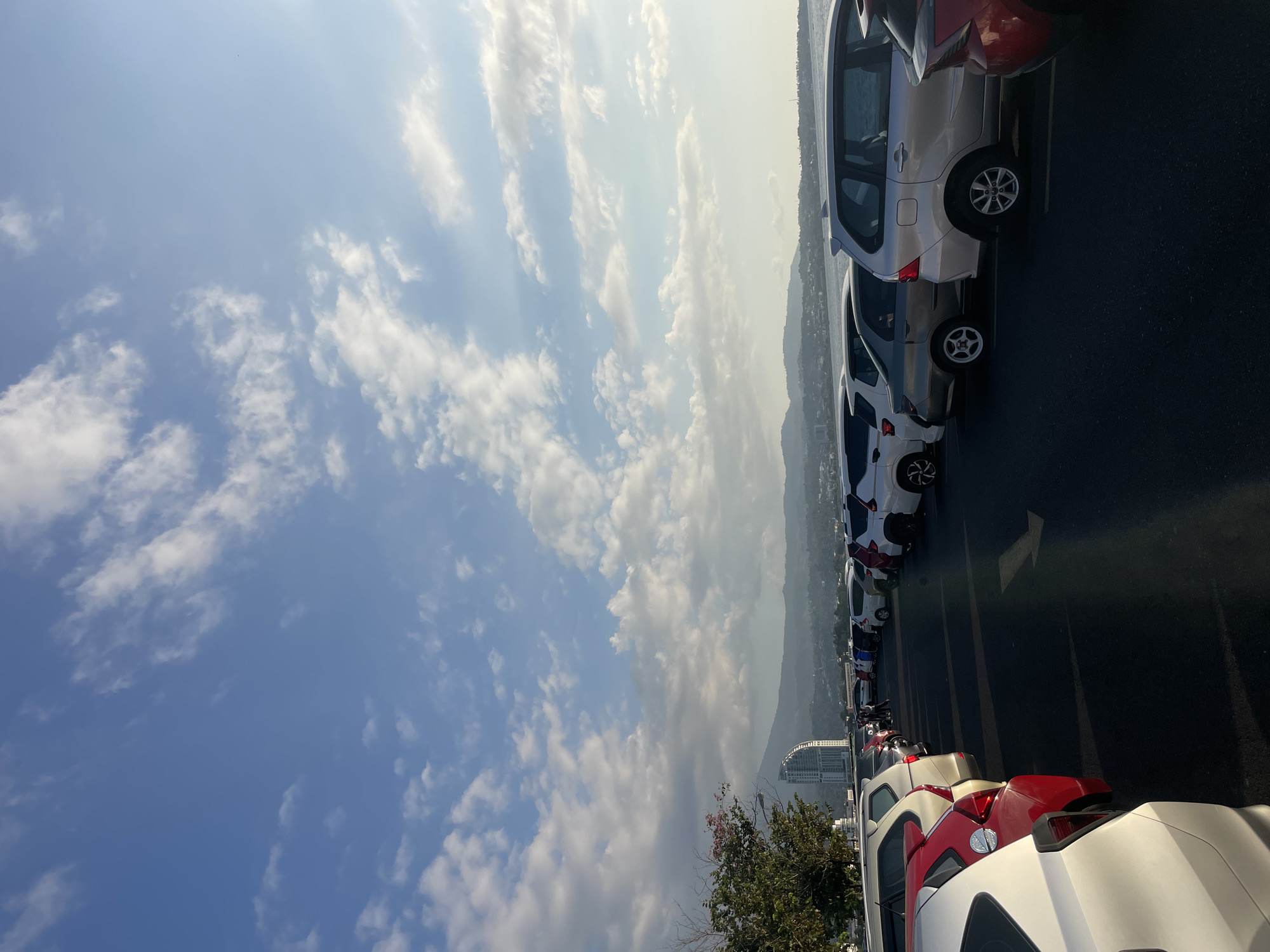 The image shows a parking lot overlooking a bay in Manado, Indonesia. Several cars are parked in a line, with a few people visible walking between them. In the background, there is a cityscape with a prominent curved building and mountains beyond. The sky is blue with scattered white clouds. The lighting suggests it is late afternoon.