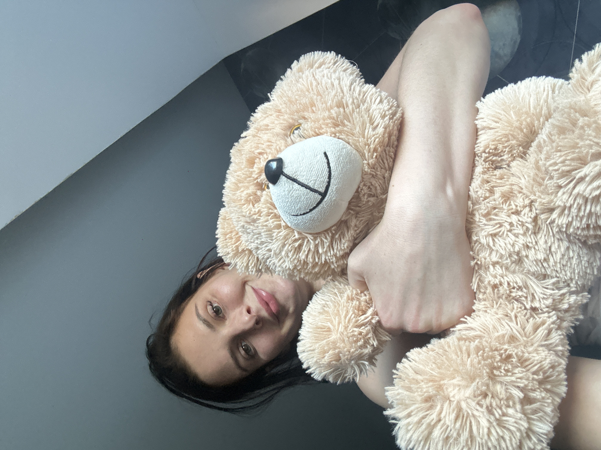 A young woman with dark hair and light green eyes is holding a large, tan teddy bear. She is smiling at the camera. The background is a muted gray wall that meets a darker wall at an angle. The lighting is dim, with a soft light source coming from the upper left. The image appears to be a selfie taken indoors.