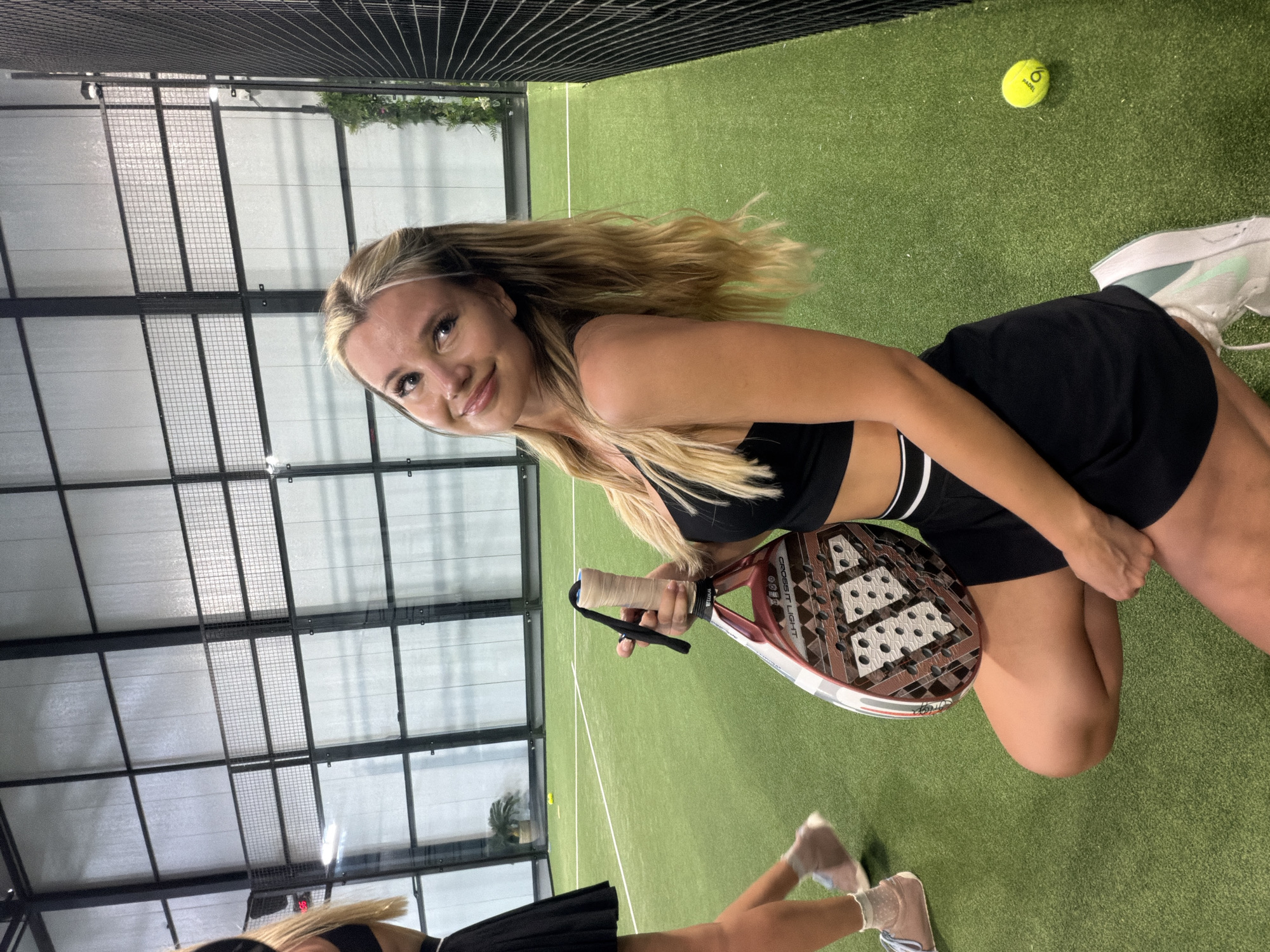 This media file shows a young woman posing for a selfie on a green court, likely a padel court, in an indoor facility. She has long blonde hair and is wearing a black sports bra and black shorts. In her right hand, she holds a padel racket with a brown grip and a white and brown patterned face. A yellow padel ball with the word "PADEL" visible is on the ground near her feet. Her left foot is also visible, wearing a white athletic shoe with light blue accents. Another person, whose legs are visible, is in the foreground, also wearing athletic shoes and a black skirt. The background shows the typical indoor court setting with a mesh fence along the top and a ceiling with a grid of lights and panels. The lighting suggests it is daytime, and there are no visible weather cues. The woman appears happy and relaxed. The location context suggests this is in Choeng Thale, Thailand.