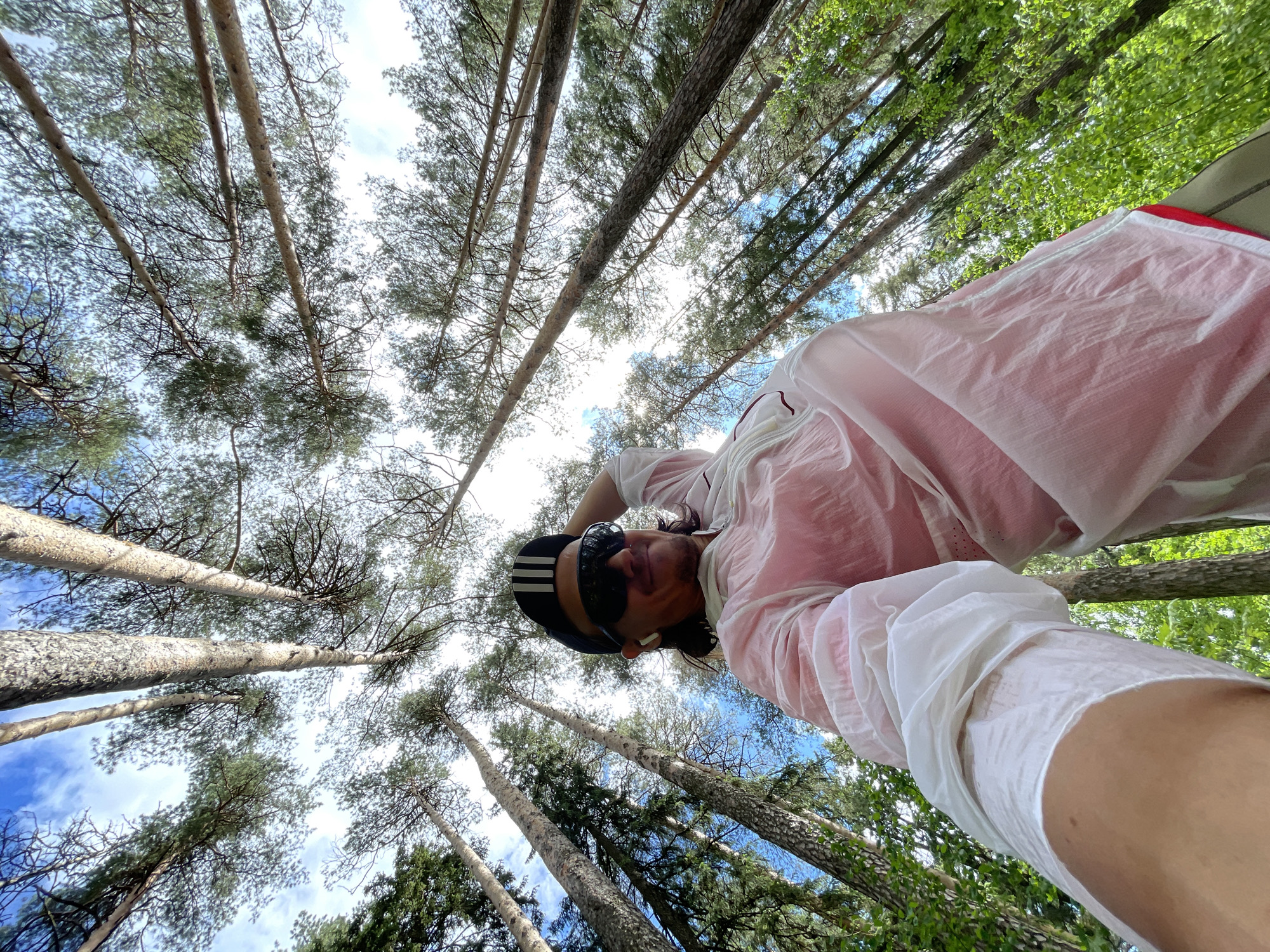 A low-angle, eye-level shot captures a man in a forest. He wears a black baseball cap with three white stripes, sunglasses, and a white long-sleeved shirt over a pink or red garment. He is looking down at the camera. Tall trees surround the man, their trunks extending upwards towards a sky with patches of blue and clouds. The forest floor is not visible. The location is not specified and therefore unknown.