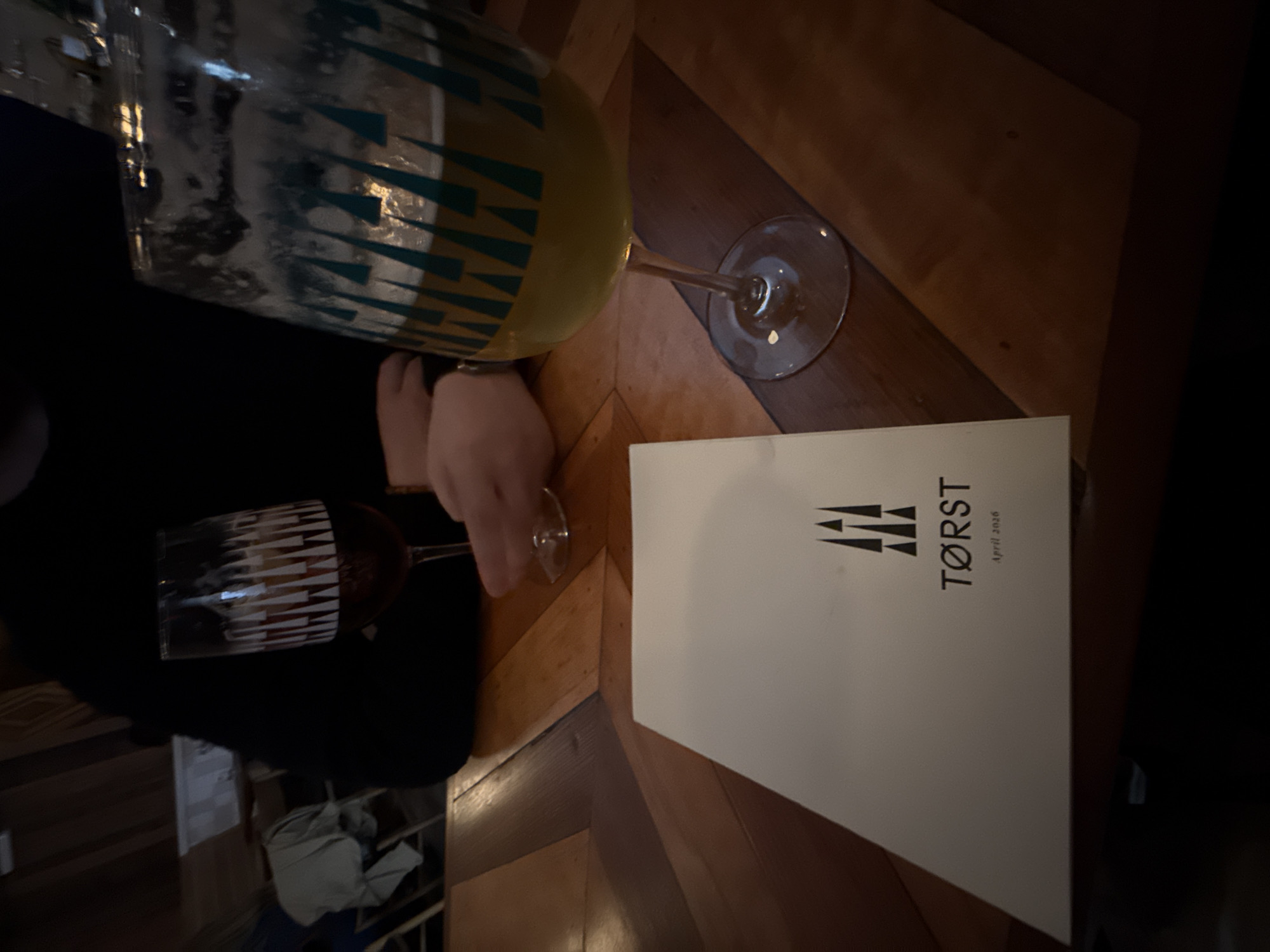 Here's a detailed description of the image:
The image shows a table setting at Tørst, a bar or restaurant in New York City, USA. A menu from Tørst with "April 2026" printed on it is on the table. A person in a black garment is holding a wine glass with red wine, and another glass with a yellow liquid is nearby on the table. The table has a geometric pattern of wood. The lighting appears dim, creating a dark ambiance. The scene conveys a social setting with beverages, possibly a dinner or drinks gathering.