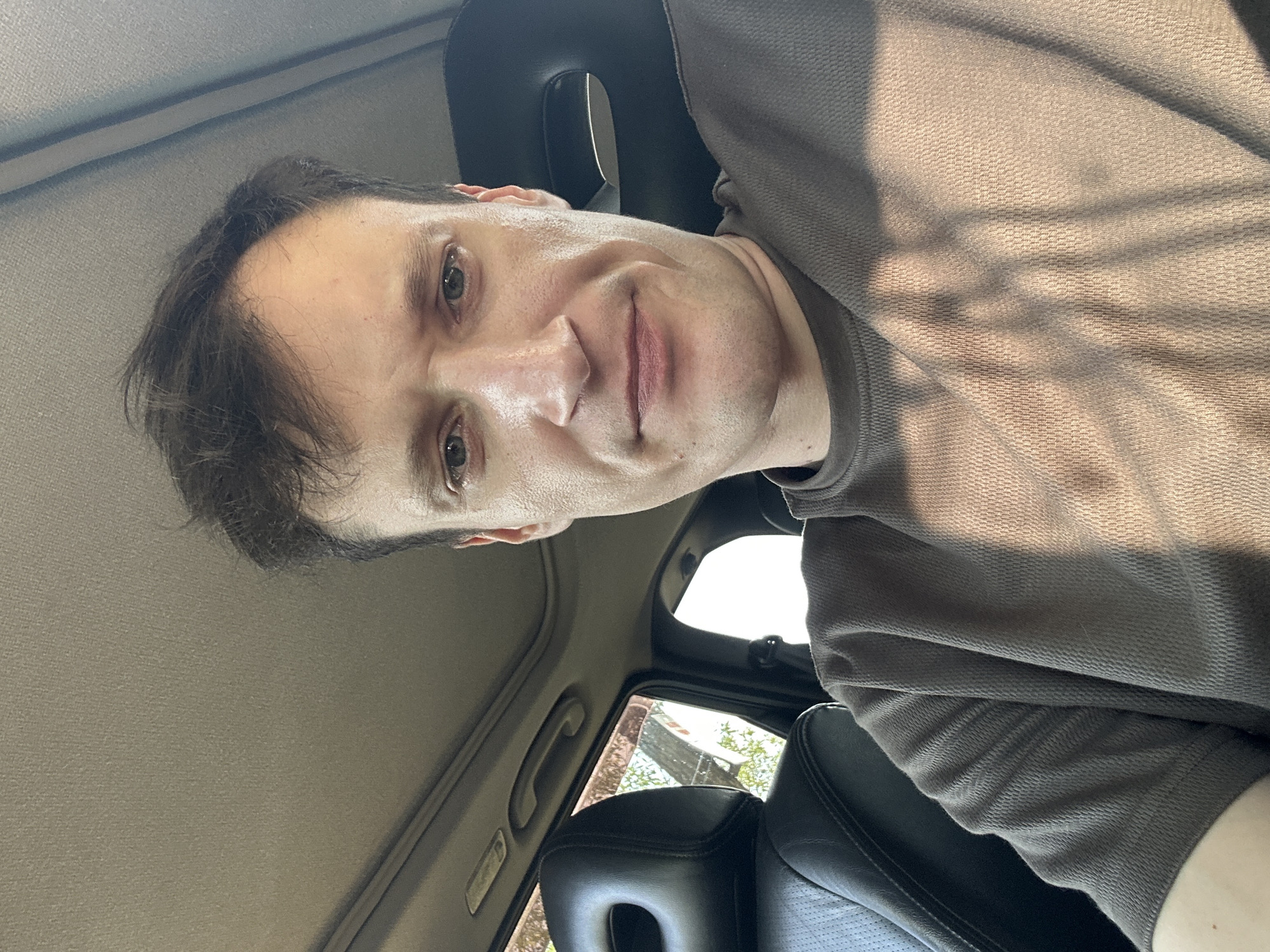 A man is seated in the driver's seat of a car, looking directly at the camera. He has short, dark brown hair and is wearing a brown t-shirt. The interior of the car is visible, including the headrest of the passenger seat, the sunroof, and the dashboard. Sunlight is streaming through the windshield, casting shadows across the man's face and t-shirt. The image appears to have been taken during the daytime. The man is located in Makiv, Ukraine.