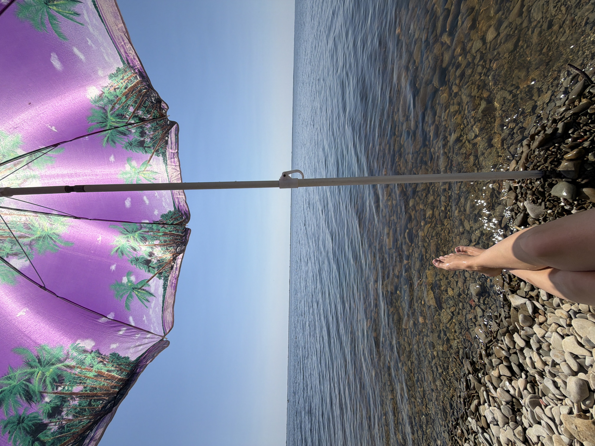 The image depicts a low-angle view of a person's legs and feet resting on a pebble beach, with a purple beach umbrella featuring a palm tree design overhead. The umbrella is supported by a white pole. The scene looks out onto a large body of water, likely the Volga River, under a clear blue sky. The water is calm with small ripples. The shore is covered with smooth, rounded pebbles of various sizes and colors. In the distance, the horizon line is clearly defined between the water and the sky, with faint dark lines that could indicate distant land or structures. The lighting suggests a sunny day. This scene is consistent with locations along the banks of the Volga River in or near the city of Kamyshin, Russia, known for its pebble beaches and riverfront.