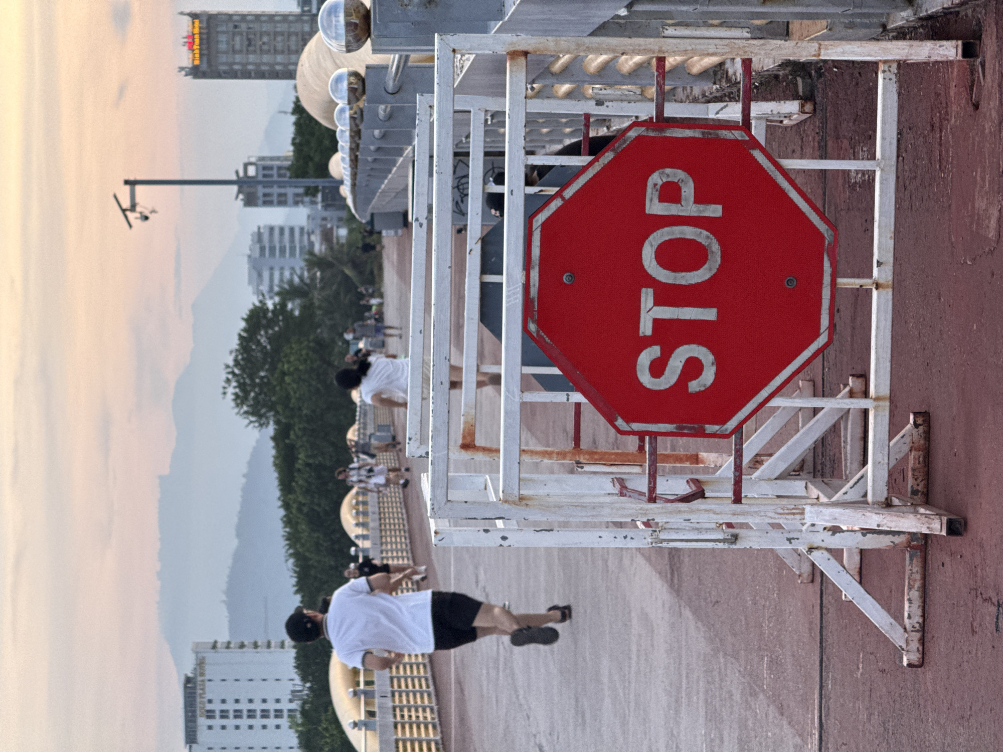 This image captures a view along a waterfront promenade in Đà Nẵng, Vietnam, as suggested by the distinct architecture of a building with the text "THE EAST COAST HOTEL" visible. The scene is bathed in the soft, warm light of either sunrise or sunset, indicating the time of day. The sky is a hazy blend of pale orange and pink hues, with distant mountains visible in the background, shrouded in a light mist.
The foreground is dominated by a red octagonal stop sign, mounted on a weathered white metal frame. The sign clearly displays the word "STOP" in bold white lettering. Behind the sign, a walkway or promenade with a reddish-brown surface stretches into the distance. Several people are visible on this walkway.
In the middle ground, a group of individuals, mostly wearing white t-shirts and dark shorts or pants, appear to be engaged in some form of physical activity, possibly exercising or jogging. Their postures suggest movement. Further into the background, more people are seen strolling or gathered.
Beyond the promenade, lush green trees line the waterfront. In the far distance, modern buildings, including the aforementioned "THE EAST COAST HOTEL" and other high-rises, punctuate the skyline, contrasting with the natural landscape of mountains and sea. The overall atmosphere seems relaxed and peaceful, with people enjoying the coastal environment. The metal structures, including the stop sign's frame and railings along the promenade, show signs of wear and tear, suggesting an outdoor environment exposed to the elements. A lamppost with a security camera is also visible in the upper left portion of the image.