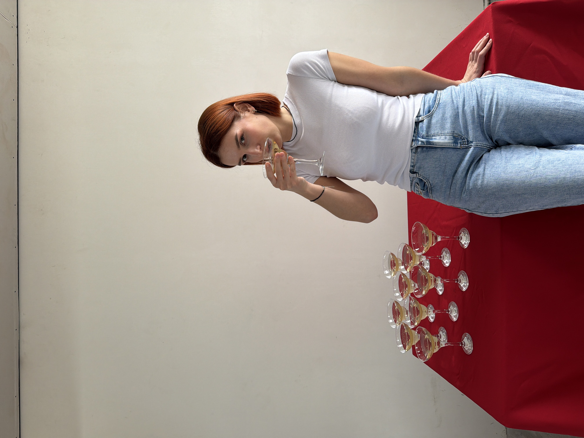 This image depicts a female subject positioned in an indoor setting. The specific location within Chaikovsky, Russia, cannot be determined from the visual information provided.
The subject is an adult female with short, reddish-brown hair, wearing a white ribbed short-sleeved t-shirt, light blue denim jeans, a thin black necklace, and a black beaded bracelet on her left wrist. A small earring is visible in her left ear. She is standing on the right side of the frame, leaning her right hip and arm against a table covered with a plain red tablecloth. Her right hand rests flat on the red surface. The subject is holding a stemmed coupe glass containing a light-colored liquid with her left hand and is in the process of drinking from it, with the glass rim at her lips. Her gaze is directed forward, towards the viewer.
On the red-covered table, to the left of the subject, nine additional coupe glasses, identical in style to the one held, are arranged in an informal cluster. Each of these glasses also contains the same light-colored liquid.
The background consists of a plain, off-white wall exhibiting minor surface blemishes. A narrow strip of a ceiling or ceiling trim, featuring small dark circular marks, is visible along the top edge of the frame. The scene is evenly illuminated.