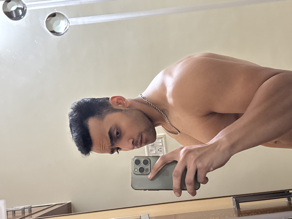 A young adult male, appearing of South Asian descent, is captured in an indoor mirror selfie in Kolkata, India. He stands shirtless, showcasing a muscular physique, and wears a silver-colored link chain necklace. His dark hair is neatly styled, and he has faint facial stubble. The subject holds an olive-green iPhone, featuring a triple-camera system, in his right hand, directing it towards his reflection in the mirror. His gaze is fixed on his mirrored image. The background, visible through the mirror, consists of a light-colored wall. An Indian-style electrical outlet with multiple switches is reflected behind the phone. A segment of a wooden frame or door is visible on the left side of the reflection. Two circular, chrome-finished fixtures are partially visible in the upper right quadrant of the reflected scene.