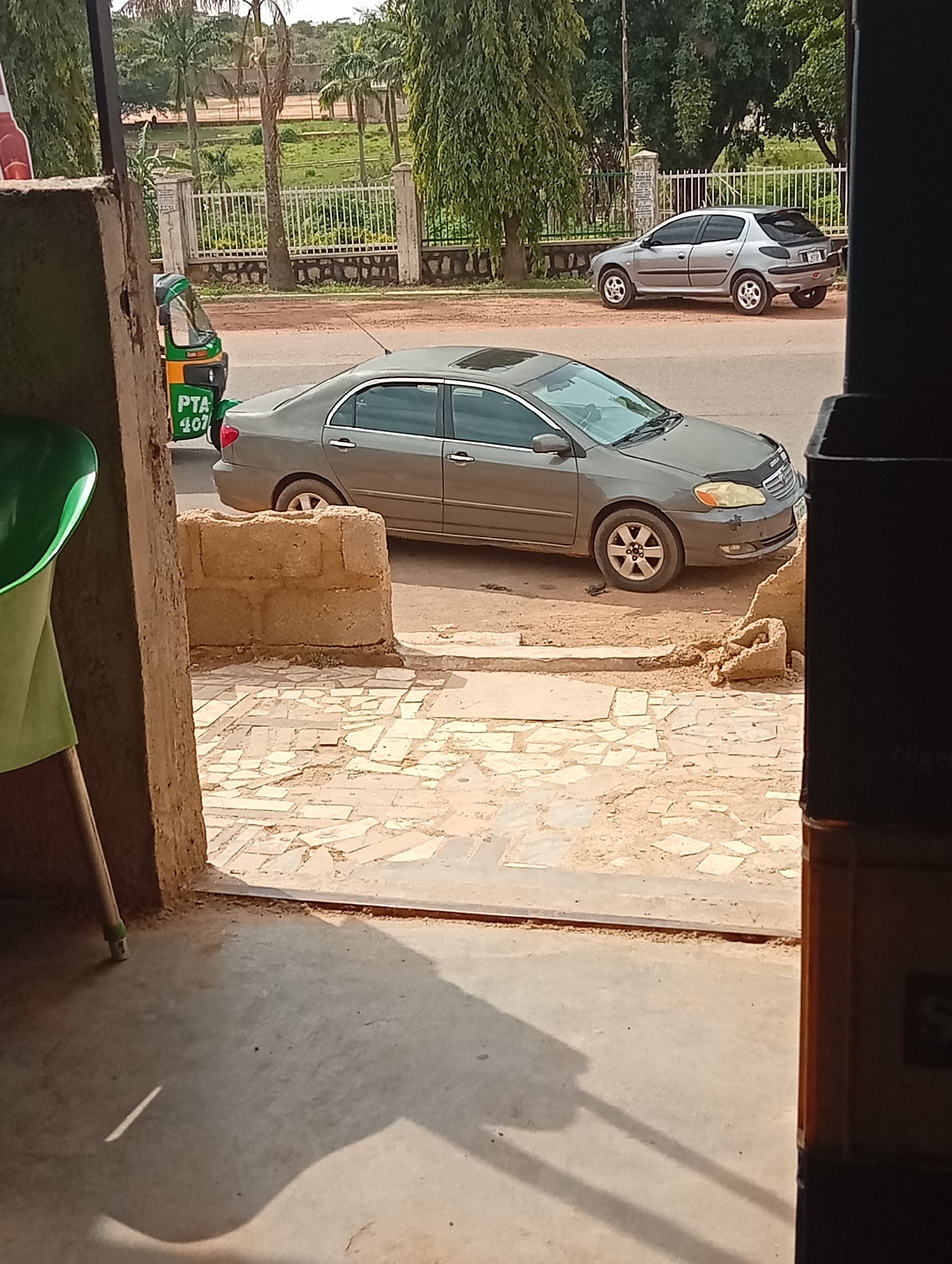 The scene, likely in Jos, Nigeria, is observed from an interior viewpoint, possibly a storefront. A gray Toyota Corolla is parked on a paved road adjacent to a pedestrian area constructed with irregular stone slabs. A small green three-wheeled vehicle (a "Keke Napep"), with "PTA 407" markings, is partially visible behind the car. Across the road, a light gray Peugeot hatchback is parked. A decorative fence runs along a grassy area with trees. A green chair is visible on the left inside the presumed storefront. Part of a brown cabinet/counter with the partial word "Niger" is visible on the right side of the image. The ground is dusty.