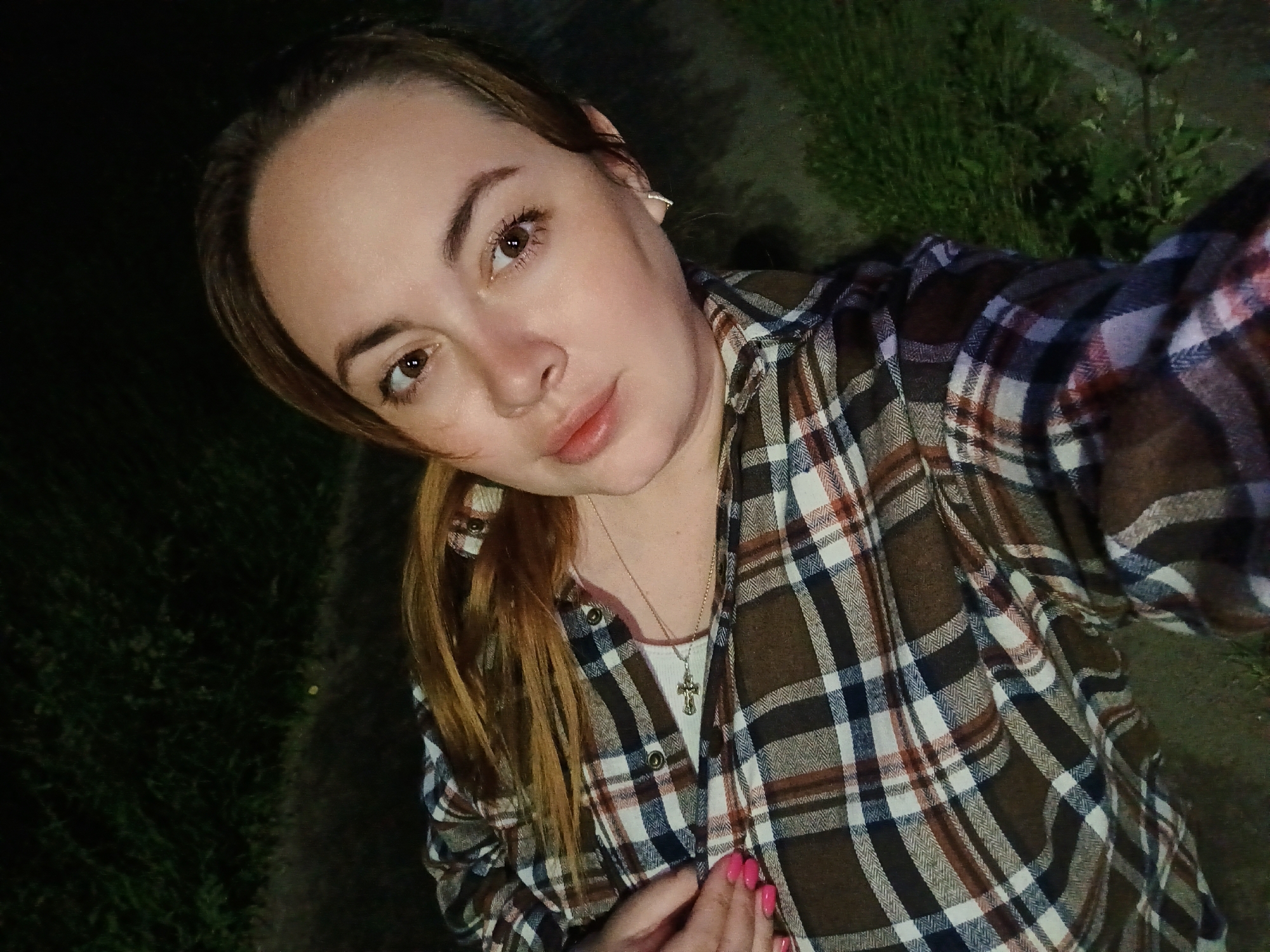 The image shows a close-up selfie of a young woman with light brown hair, tied back in a ponytail. She is wearing a plaid shirt over a white tank top, and a silver cross necklace. Her fingernails are painted bright pink. The background is dark and out of focus, but appears to be an outdoor setting with grass and a path. The woman's facial expression is neutral, and she is looking directly at the camera. The lighting suggests the photo was taken at night or in low light conditions. There are no discernible landmarks to indicate a specific location within the city of Chaikovsky, Russia.