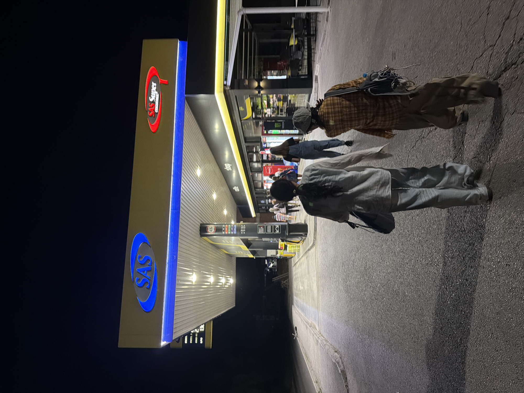 The image depicts a night scene at a SAS Super gas station in Mali Zvornik Municipality, Serbia. Two women are walking away from the camera on the asphalt surface of the gas station. They are walking towards a brightly lit building that appears to be a convenience store or shop, attached to the gas station. The building has a yellow and black facade with illuminated signage that reads "cafe" and "shop." Inside the shop, shelves stocked with various items are visible through the glass windows. The gas station canopy is illuminated with bright lights, and the SAS logo is prominently displayed in blue and red. Several cars are parked at the gas pumps. The overall environment is well-lit, suggesting the gas station is open for business at night.