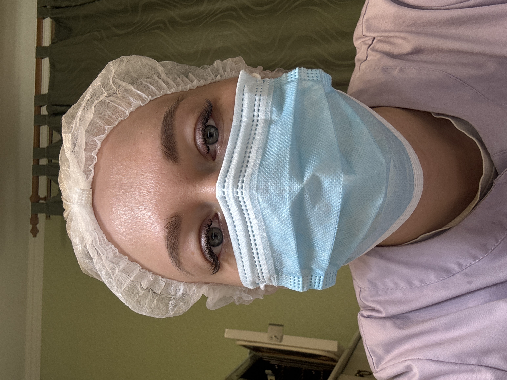 A person wearing a light blue surgical mask and a white hairnet looks directly at the camera. They are also wearing a light purple collared shirt. The background appears to be an examination room setting. The location is Ферма, Russia.