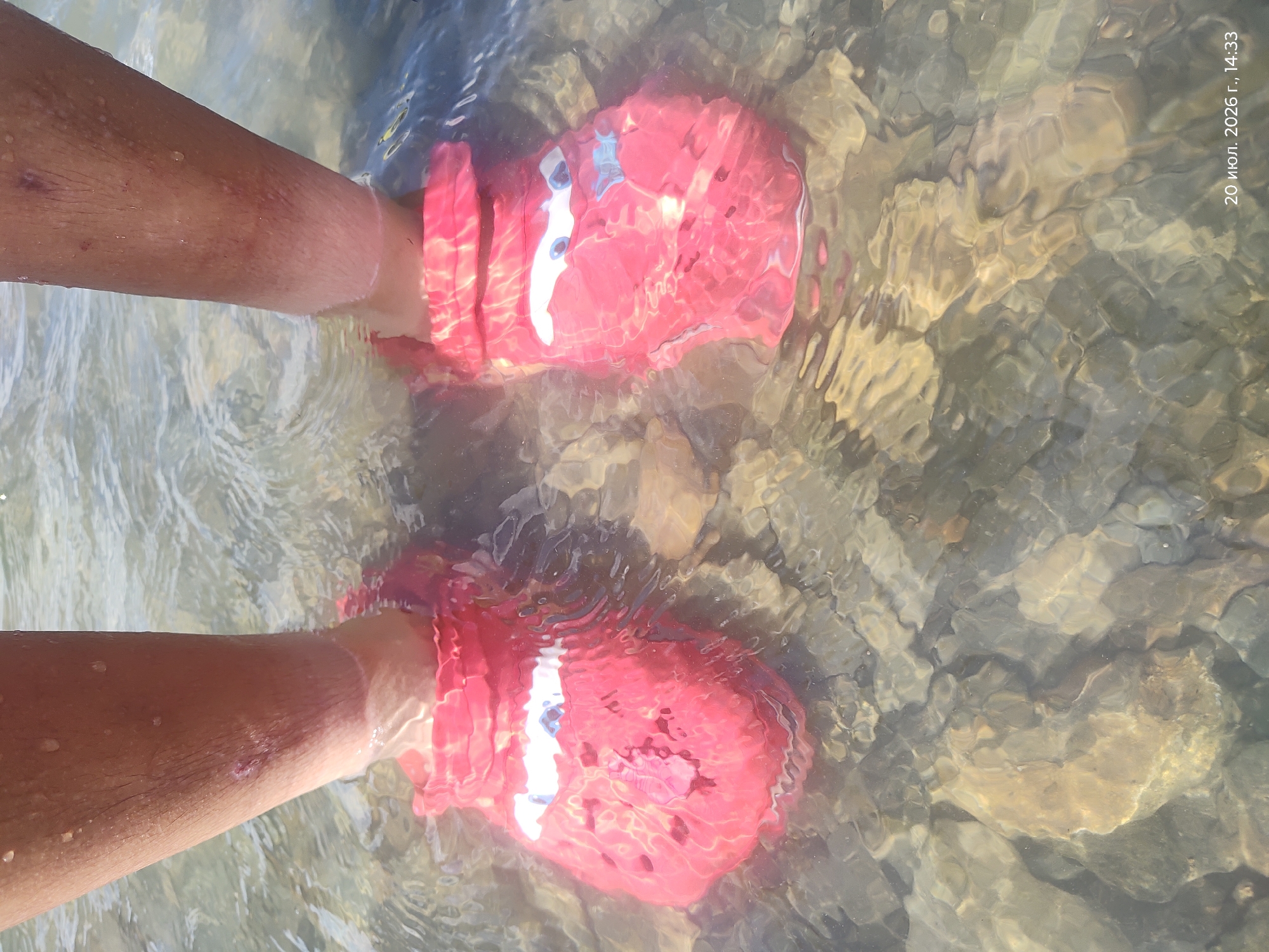 Two bare legs wearing bright pink crocs with a lightning McQueen design are submerged in clear, shallow water. The crocs are partially covered by the water, which distorts their appearance. The water is so clear that the rocky riverbed beneath is visible, with stones of various sizes and colors. The legs are positioned close together, and the focus is on the footwear and the interaction with the water. The scene appears to be in a natural outdoor environment, possibly a river or a shallow stream. The timestamp "20 июля. 2026 г., 14:33" is visible in the bottom right corner of the image, indicating the date and time the photograph was taken.