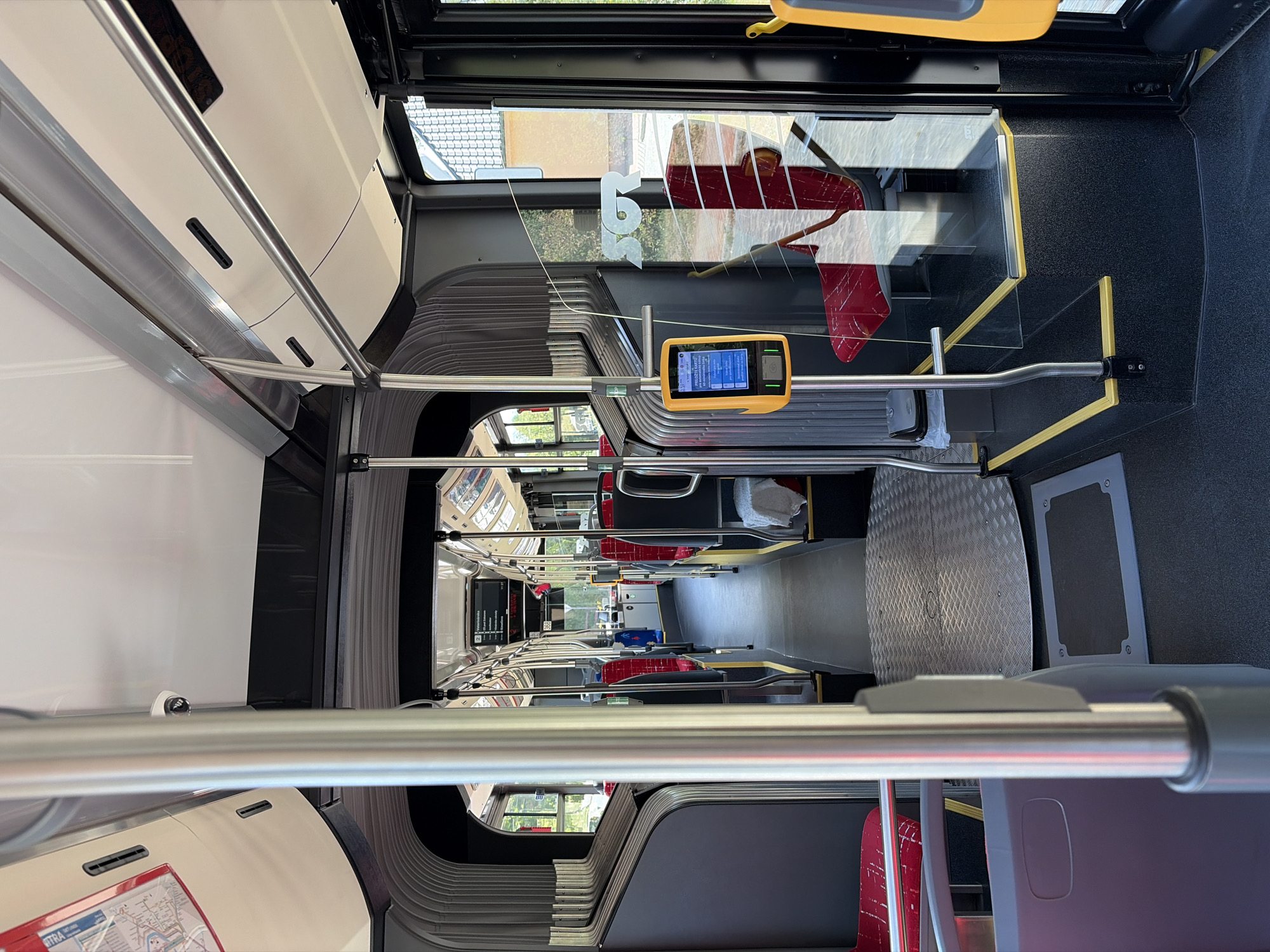 The image shows the interior of a modern public transport bus, likely a trolleybus, in Nitra, Slovakia. The perspective is from the aisle of the bus, looking towards the rear. The bus features red seating with silver frames and a dark grey interior. Poles and handrails are visible throughout, indicating passenger safety features. A ticket validator is mounted on a pole in the foreground, displaying information on its screen. The floor is a dark grey rubberized material with yellow safety markings. The ceiling is white with integrated lighting. In the background, digital route information displays are visible, showing destinations such as "Voderady" and "Jur nad Hronom". The overall environment suggests a clean and functional public transportation setting.