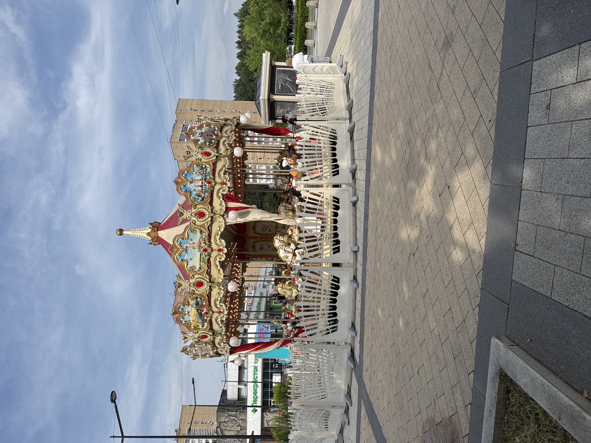 The image shows a carousel situated in an urban plaza in Zelenograd, Russia. The carousel, adorned with red and gold decorations, features a central ornate structure with painted panels depicting various scenes. Numerous white horses with golden accents are arranged around the carousel's perimeter. A white, decorative fence encloses the carousel, with some sections appearing to be movable barriers. In the background, there are multi-story buildings, including a building with a green sign that says "Перекресток" (Perekrestok), a Russian supermarket chain. Trees line the background, and a clear sky with scattered clouds is visible above. Two individuals are present near the carousel: one person, likely a woman, stands near the carousel entrance, and another person is walking away from the carousel. The plaza is paved with interlocking stone tiles. Streetlights and utility poles are also visible in the scene.