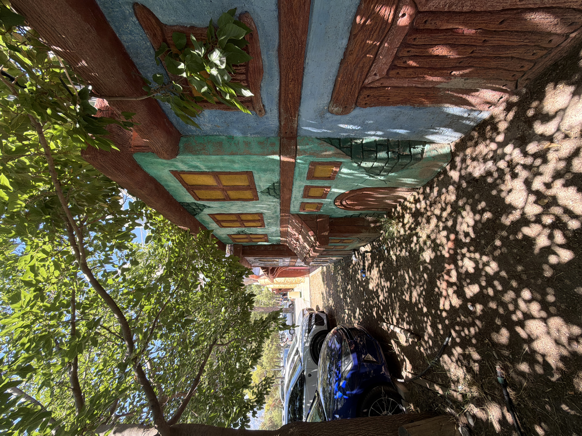 The image depicts a colorful, stylized building facade, painted in shades of blue, teal, and brown, with accents of yellow for the window panes. The building appears to be constructed to resemble older architecture, with textured surfaces and faux wooden elements. In the foreground, a blue car and a white car are parked along a gravel path, partially shaded by a large tree with lush green leaves overhead. The ground is littered with dry leaves and small debris, with dappled sunlight casting shadows from the tree. In the background, more trees and what appears to be a street with other vehicles and pedestrians can be seen. The scene suggests an outdoor setting, possibly a pedestrian walkway or a street with shops, in a location with a Mediterranean climate, consistent with Kemer, Turkey.