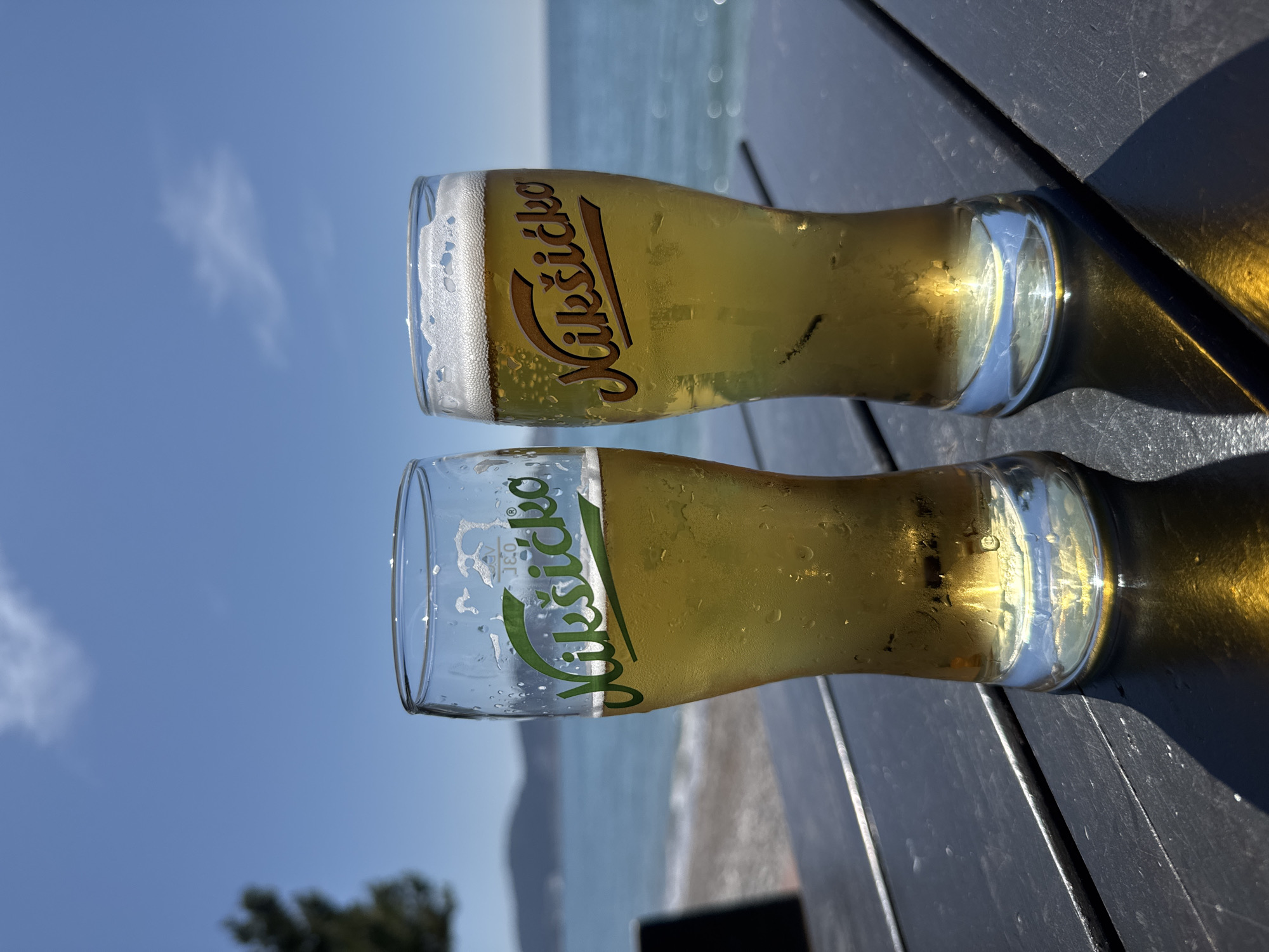 Two glasses of "Nikšićko" beer are sitting on a dark-colored table in an outdoor setting. The beach and sea are visible in the background. The scene is likely in Bar, Montenegro, due to the prominent branding of the Nikšićko beer, a popular Montenegrin beer brand. There are no people visible in the image.