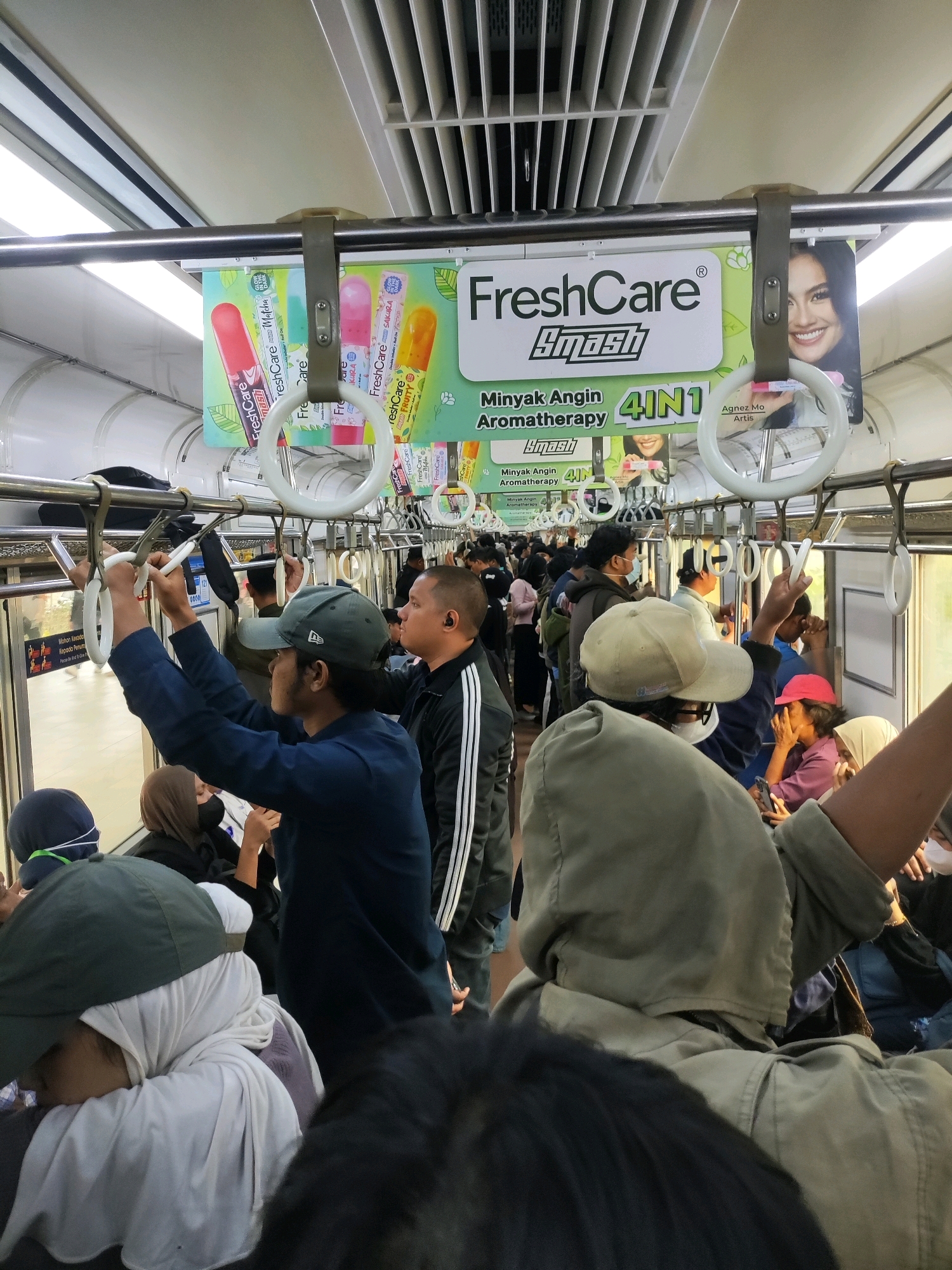 The image depicts the interior of a crowded train carriage, likely on a commuter line, with the environment suggesting it is within Bekasi, Indonesia, due to common transportation seen in the region. The perspective is from a passenger standing amidst a throng of people, looking upwards and slightly forward.
In the foreground, the backs of several passengers are visible. One individual in a dark blue jacket and a dark green baseball cap is holding onto a white handrail strap. To their right, another person in a black jacket with white stripes on the sleeves is also holding a handrail. Further to the right, a person wearing a pink cap is partially visible. In the immediate foreground, a person wearing a light-colored hooded jacket and a tan baseball cap is prominent, their back to the camera. Another passenger in a white head covering and a dark blue outer garment is seated or leaning forward, their face obscured.
The middle ground is filled with a dense crowd of passengers standing and holding onto the numerous handrails and poles that line the carriage. Faces are largely indistinct due to the low lighting and the density of the crowd, but various headwear, including hijabs and caps, are visible.
The background shows the upper interior of the train carriage, including the ceiling with ventilation grilles and fluorescent lighting. A large advertisement banner is suspended above the handrails, featuring the "FreshCare Smash" logo, product imagery of aromatherapy roll-ons, and text in Indonesian, including "Minyak Angin Aromatherapy 4IN1". The face of a woman, identified as "Agnez Mo Artis," is also part of the advertisement. The overall impression is one of a busy, public transportation setting during what appears to be peak travel time.
