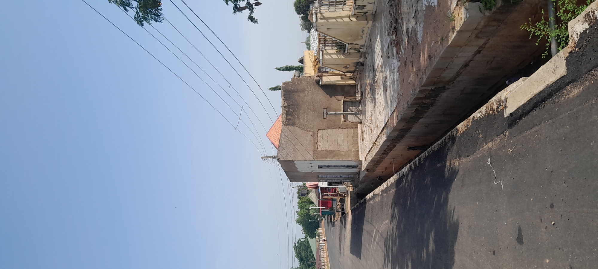 The image depicts a street scene in Jalingo, Nigeria, on a clear day under a bright blue sky. The foreground features an asphalt road with a drainage ditch running alongside it. On the right side of the road, a beige, unfinished concrete building with a white door is prominent. Adjacent to this building is a raised, paved area with a small wooden table and stool. Further back, a yellow wall with a gate topped with barbed wire encloses a property. In the background, along the left side of the road, are several small shops with colorful awnings, including one with a red and white striped roof and another with a green roof. Trees and other buildings are visible further down the street. Power lines crisscross the sky above the scene. There are no people or animals clearly visible in the image. The overall impression is one of a developing urban area.