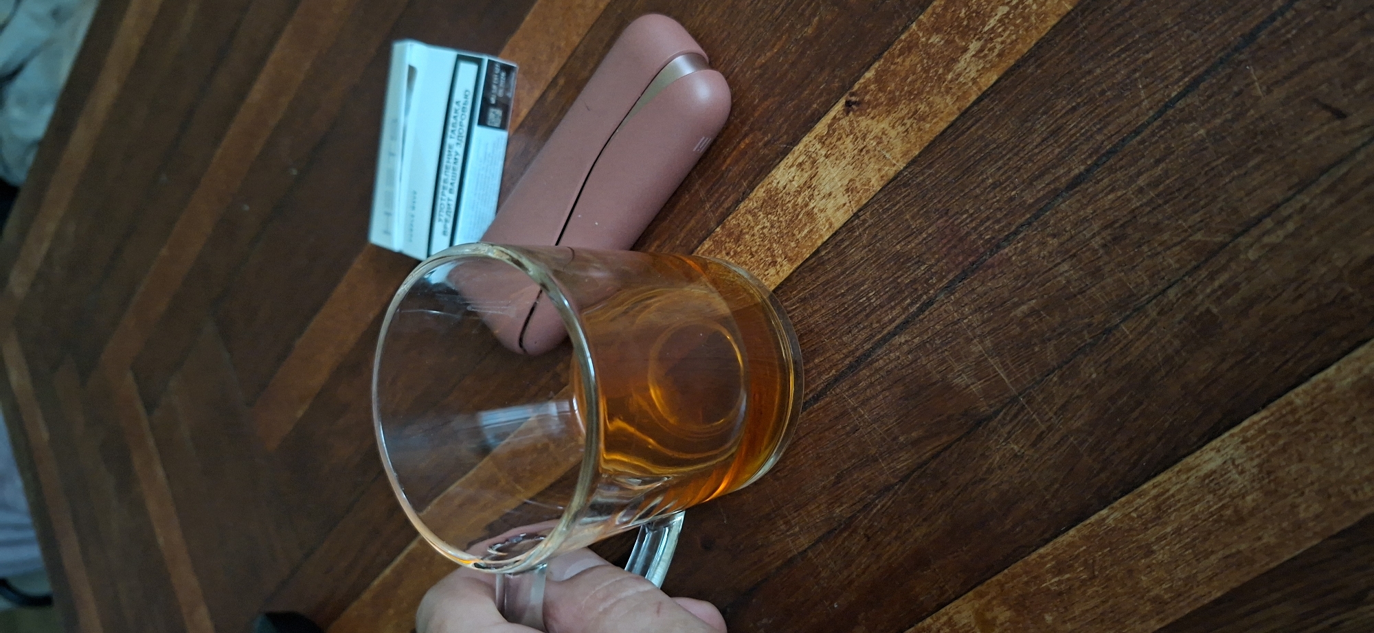 A close-up, eye-level shot shows a transparent glass mug filled with amber-colored liquid, resting on a dark wooden table with a herringbone pattern. A hand is visible on the left side of the mug, holding it. To the right of the mug, a pink electronic smoking device and a white pack of cigarettes are placed on the table. The background is blurred, showing more of the wooden table. The scene appears to be indoors. The image does not contain information about Shakhtarsk, Ukraine.