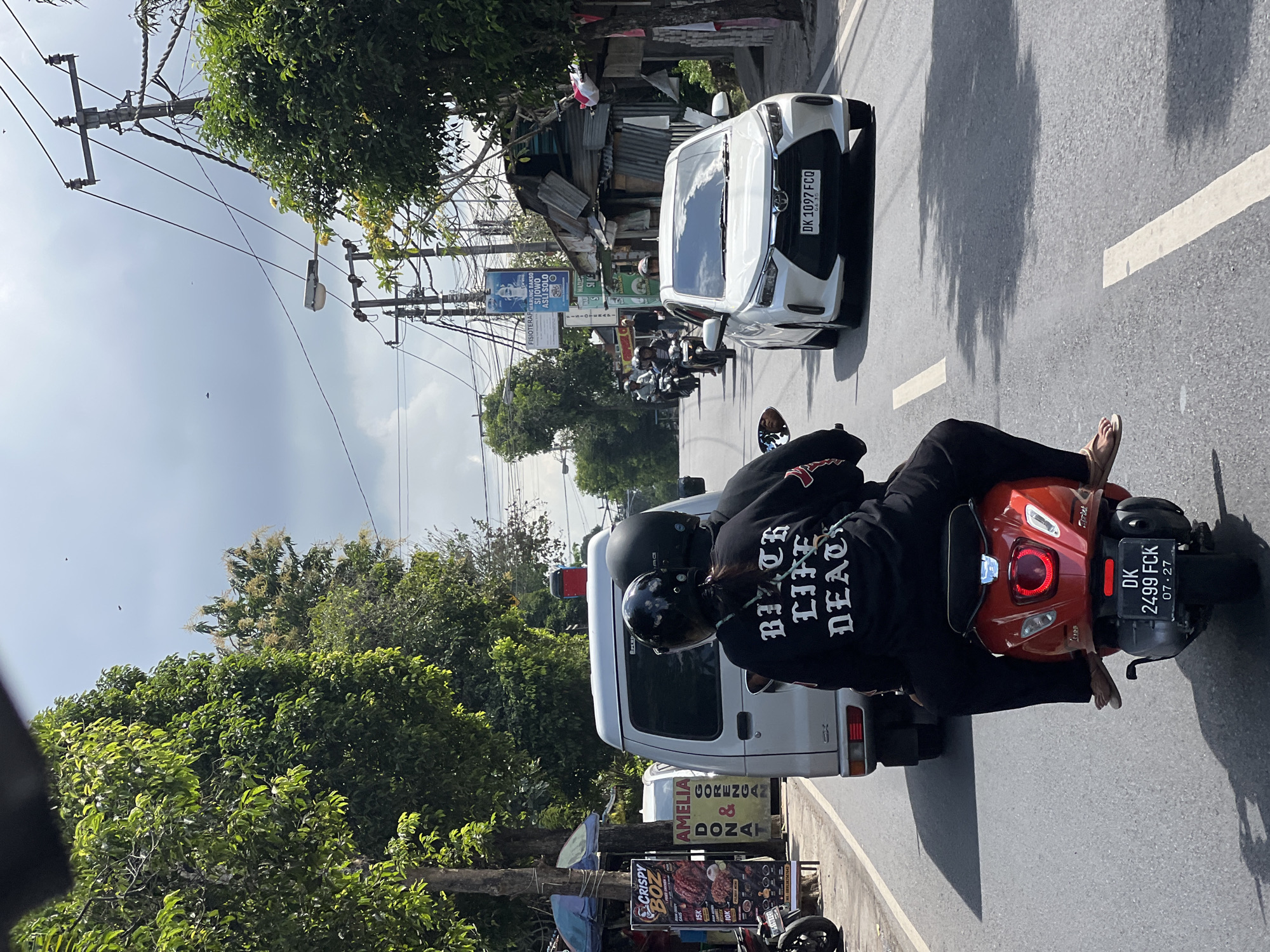 On a cloudy day in Abiansemal, Indonesia, two people are riding on a red scooter on a paved road. The rider in the back, a woman, is wearing a black sweatshirt with white lettering that reads "BITCH LIFE DEATH". The driver, also wearing a black helmet, is positioned in front of her. Behind them, a white van and a white car are also on the road. The environment is a street with trees, utility poles, and buildings lining the sides. A street sign indicating "FISIOTERAPI SI JOWO ASLI SOLO" is visible on the right side of the road. The license plate of the car reads "DK 1097 FCQ". The license plate of the scooter reads "DK 2499 FCK".