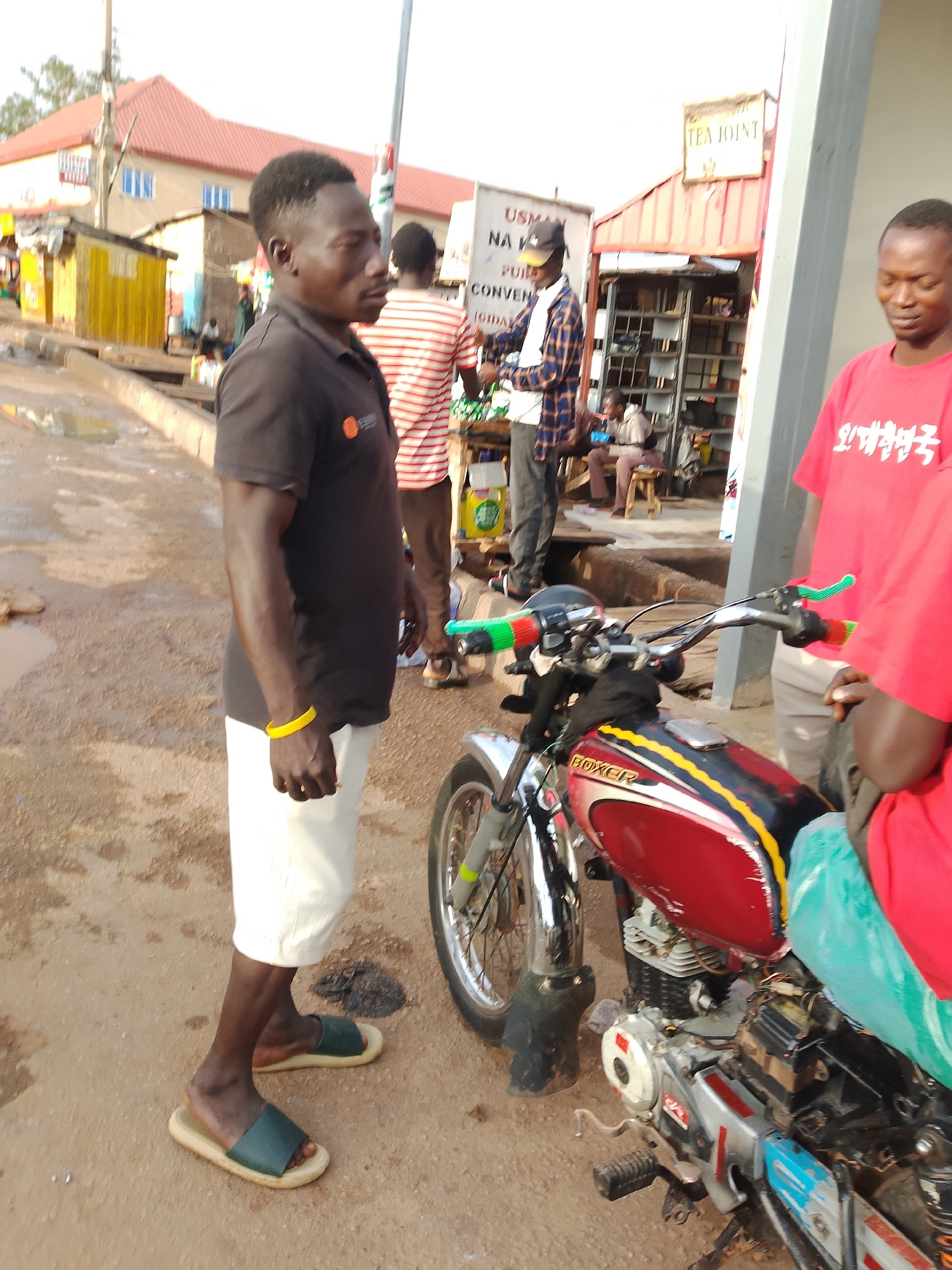This media file depicts an outdoor scene in Jos, Nigeria. Three men are visible, two of whom are the primary subjects. One man, wearing a black polo shirt and white shorts, stands to the left, facing away from the camera and looking to his right. He has a yellow bracelet on his left wrist and wears green and beige slide sandals. To his right, a red motorcycle is parked. A second man, wearing a red t-shirt with Korean text, stands to the right of the motorcycle, facing towards it and partially obscured by the motorcycle. A third man, wearing a red and white striped shirt, is visible in the background, near a stall.
The setting appears to be a street or market area. Several stalls and buildings line the background, including one with a sign that reads "TEA JOINT" and another with "USMAN NA". The ground is uneven and appears wet in some areas, suggesting recent rain. The lighting suggests it is daytime, likely late afternoon, with the sun casting shadows. The overall mood seems casual, with the men appearing to be engaged in conversation or a transaction. The motorcycle is a prominent object, painted red with yellow and black accents, and the word "BOXER" is visible on its fuel tank. The image captures a slice of everyday life in a Nigerian urban environment.