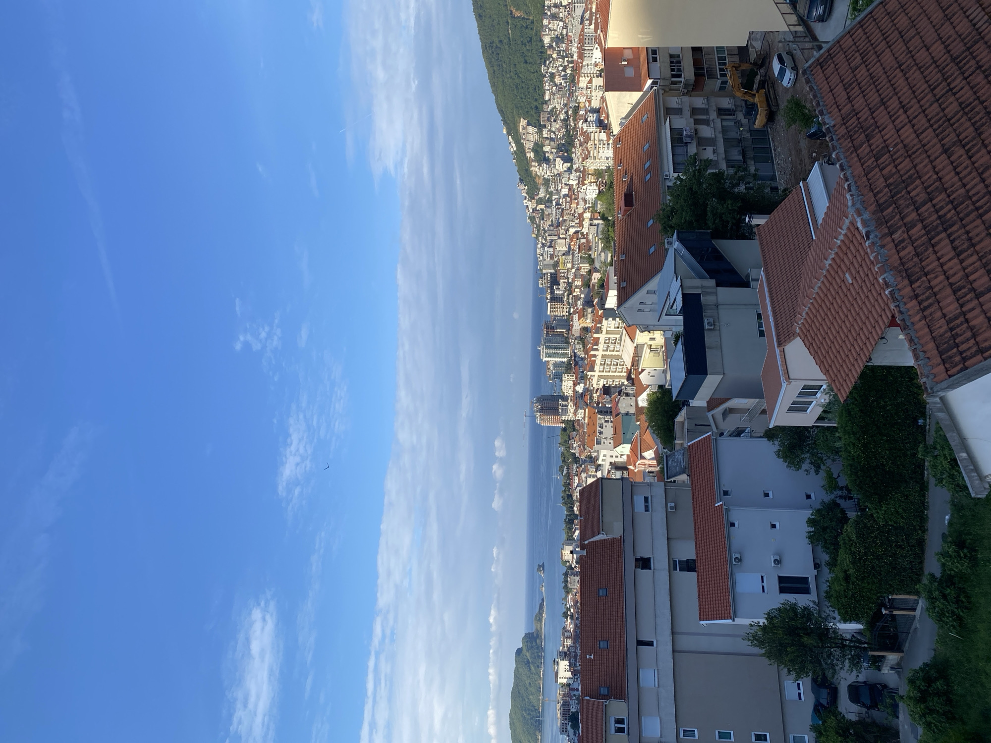 The image depicts a view in Budva, Montenegro, showcasing the cityscape along the Adriatic coast. The left side of the image is dominated by a clear blue sky with some streaky clouds. The center shows the Adriatic Sea with islands and the coastline, densely packed with buildings. The right side of the image reveals a residential area with buildings of varying heights and architectural styles, many featuring red-tiled roofs. An excavator is visible behind buildings. Trees and other greenery are interspersed among the buildings.