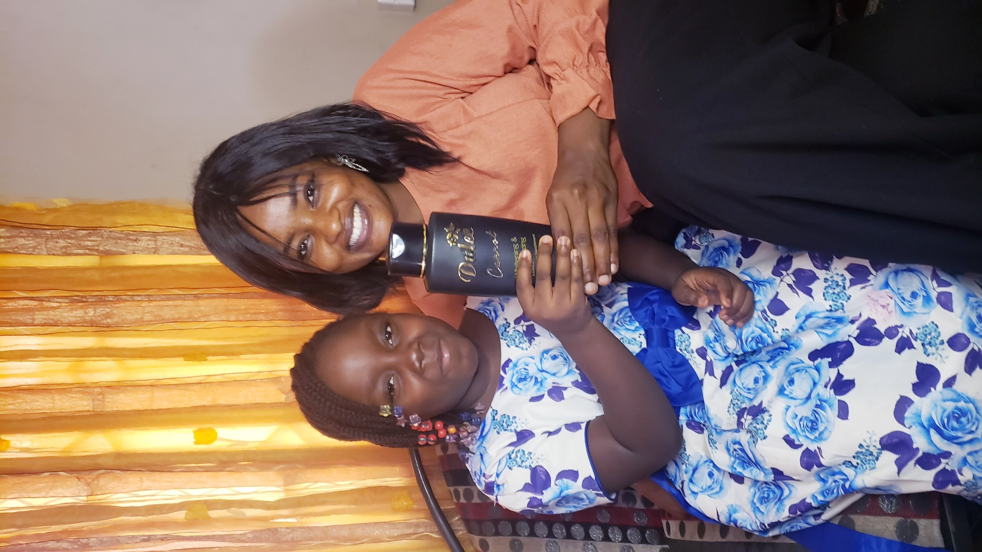 The image depicts two dark-skinned individuals, likely a mother and daughter, posing indoors. The woman, in an orange long-sleeved blouse and black pants, smiles at the camera while holding a black bottle labeled "Dulee Carrot" with the daughter. The child wears a white dress adorned with large blue floral patterns and a blue sash. She also has beads in her braided hair. The background consists of a beige patterned curtain and a couch with a patterned seat. The location is inferred to be in Narayi, Nigeria.