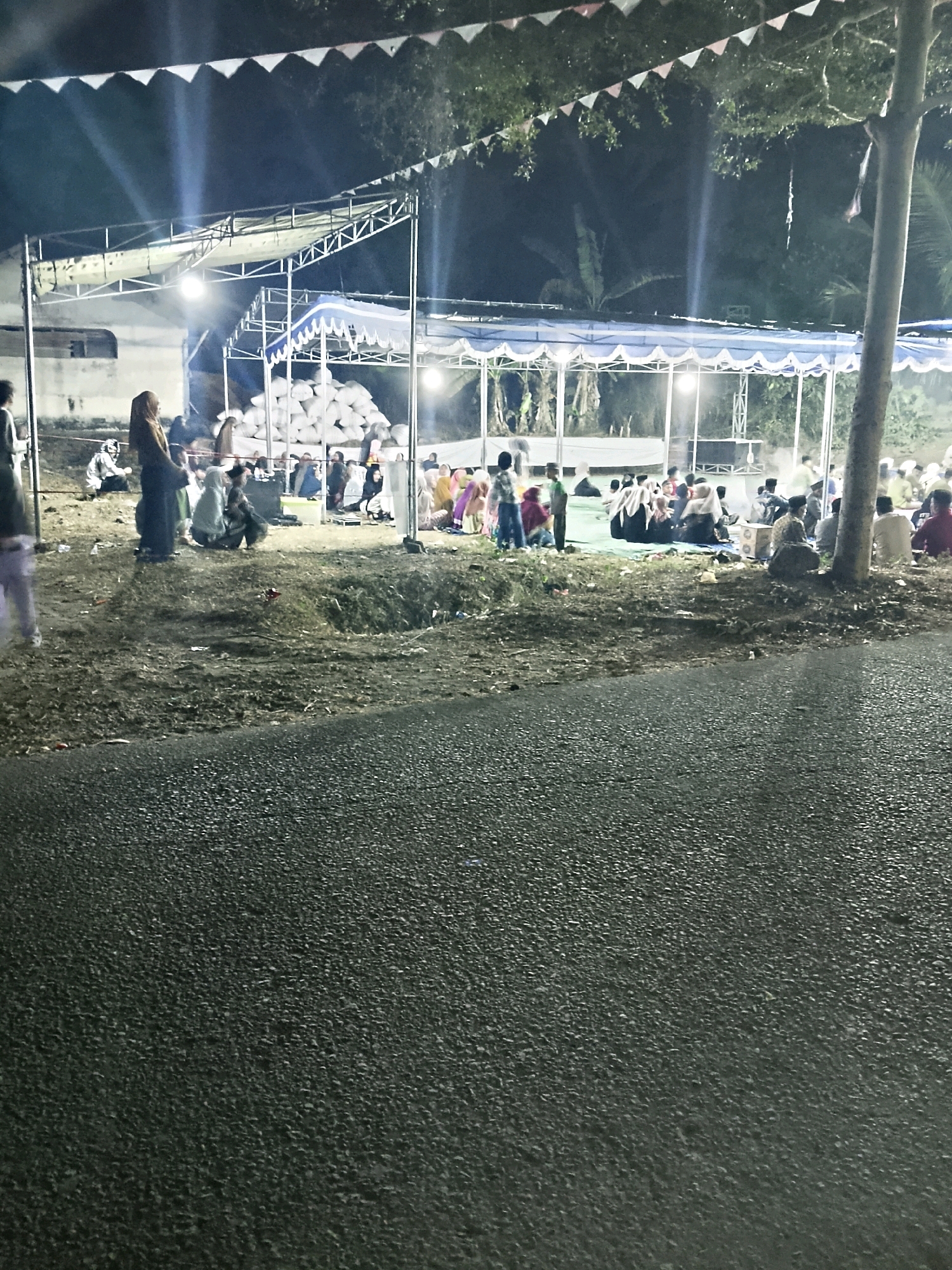 In an outdoor nighttime scene, a group of people are gathered under a temporary tent structure with lights. The ground appears to be a mix of dirt, gravel, and some grass. A paved surface, likely a road, is in the foreground and is wet, suggesting recent rain or a damp environment. The tent is supported by a metal frame and draped with fabric. A large pile of white, bag-like objects is visible behind the tent. Several individuals are seated on the ground, while others stand. The overall atmosphere is dimly lit, with artificial lights illuminating the gathering area. Trees and foliage are visible in the background, indicating a semi-rural or natural setting. The presence of a road and a building structure suggests the area may be on the outskirts of a settlement or an event ground.