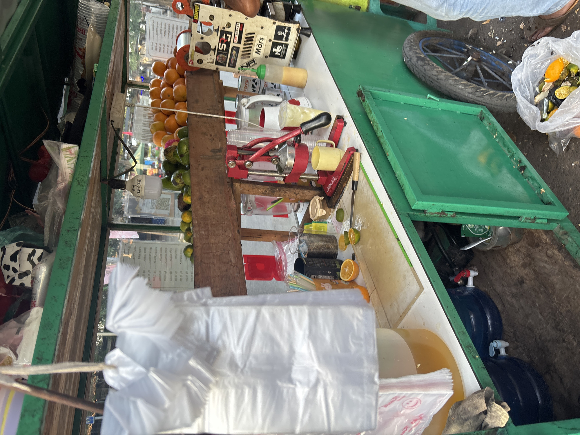 This image depicts a juice cart likely located in the city of Cirebon, Indonesia, based on the language visible on some signage. The cart is predominantly green and appears to be operating outdoors, possibly on a street or in a market area.
In the foreground, a white plastic bag filled with what appears to be citrus peels and some whole fruit is placed on the ground. To the right, a person's legs, wearing light blue jeans and brown sandals, are visible.
The cart itself is equipped for preparing and serving juice. On the lower shelf, two large blue water cooler jugs with spigots are visible, suggesting a water supply for the cart. A metal bucket with a spoon and a green package is also present.
The main working surface of the cart is a white countertop. On it, there is a red manual juicer, a yellow mug, a cutting board with sliced oranges and limes, and a knife. Various containers are also present, including a clear cup with a straw, a pitcher, and a bottle with a green cap containing a yellow liquid. To the right of the juicer, a white board with several stickers and text, including the word "Mars," is attached.
The upper shelf of the cart displays a variety of items, including stacked oranges, avocados, and what appear to be plastic bags. Further back, additional items like bottles and containers are visible. The cart also has a metal frame and a wheel visible on the right side. The background shows a blurred outdoor environment, with some stalls or buildings in the distance and trees.
