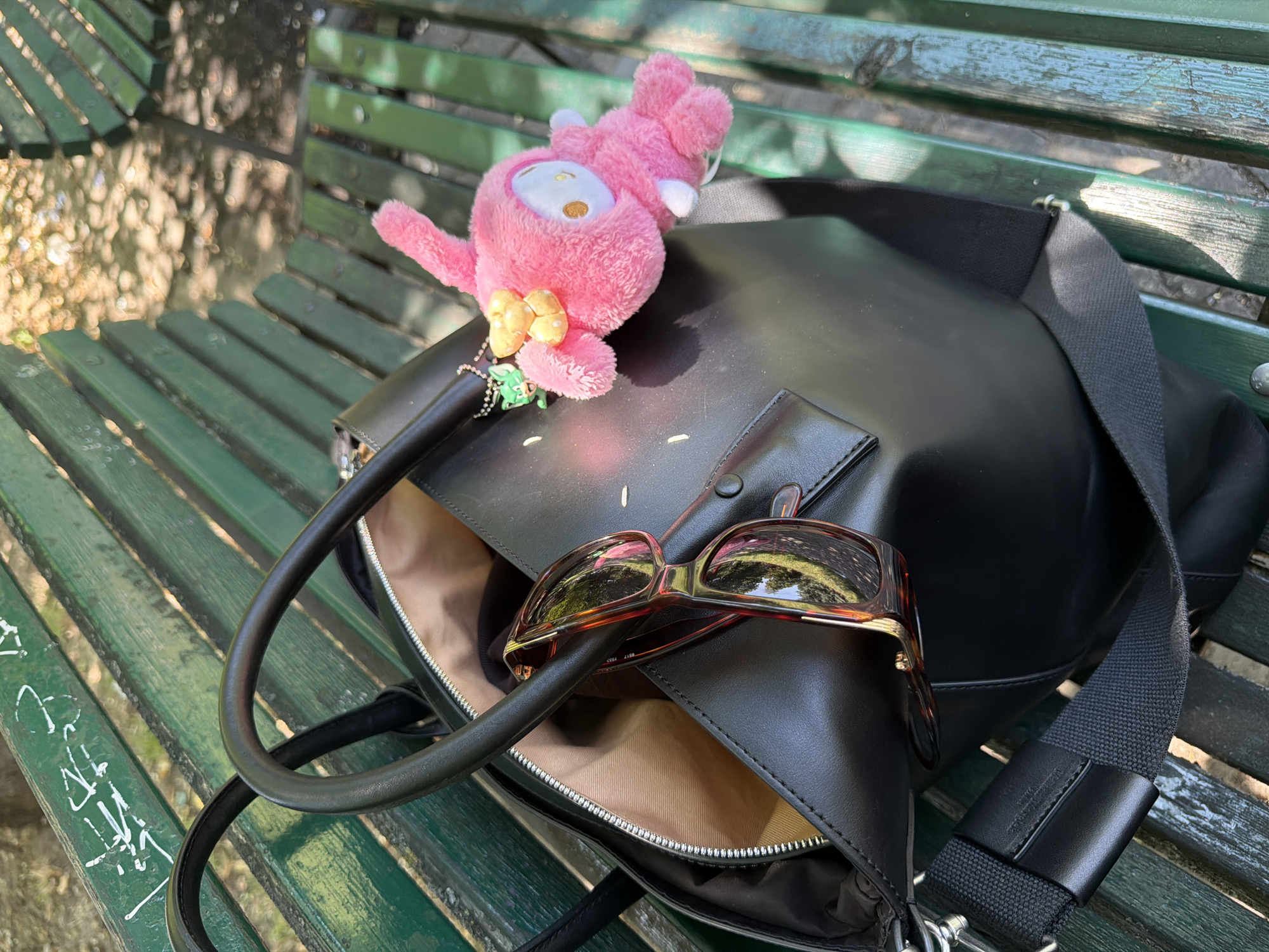 An open black leather tote bag rests on a green slatted park bench. A pair of tortoiseshell sunglasses is placed on top of the bag. A pink plush toy with yellow polka dot ears and a yellow bow hangs from a keychain attached to the bag, accompanied by a small green charm. The bench is situated in a park setting, with dappled sunlight filtering through trees. The bench has graffiti scrawled on its surface. The image captures a moment of rest on a sunny day, suggesting a casual outing in an urban park. The location is identified as Milan, Italy.
