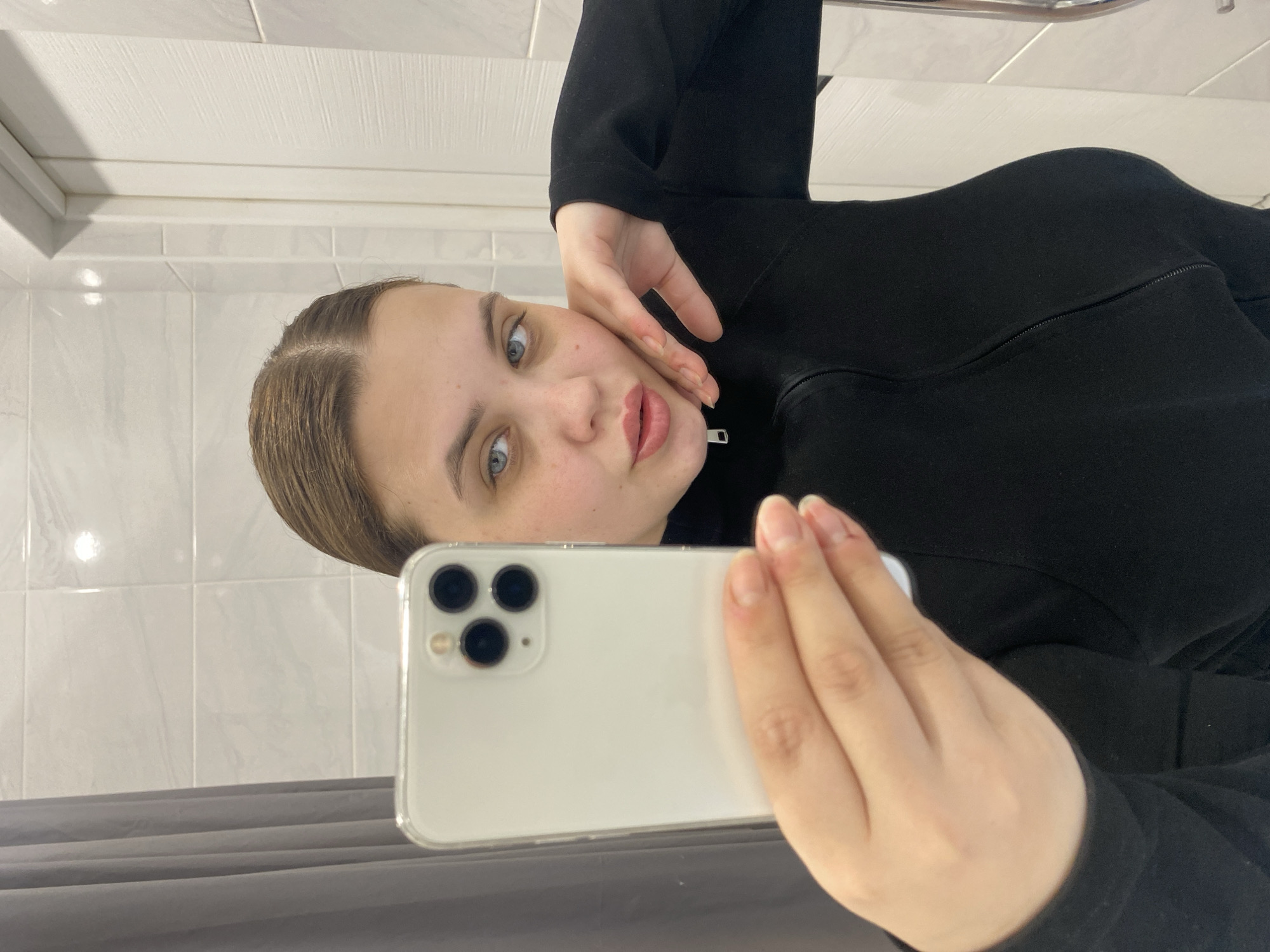 A woman is taking a selfie in a bathroom. She is holding a white smartphone with a triple-lens camera. The bathroom has white tile walls and a gray curtain rod. The woman is wearing a black zip-up jacket. Her left hand is near her face.
I have no information about the location being in the city of Chernushka, Russia.