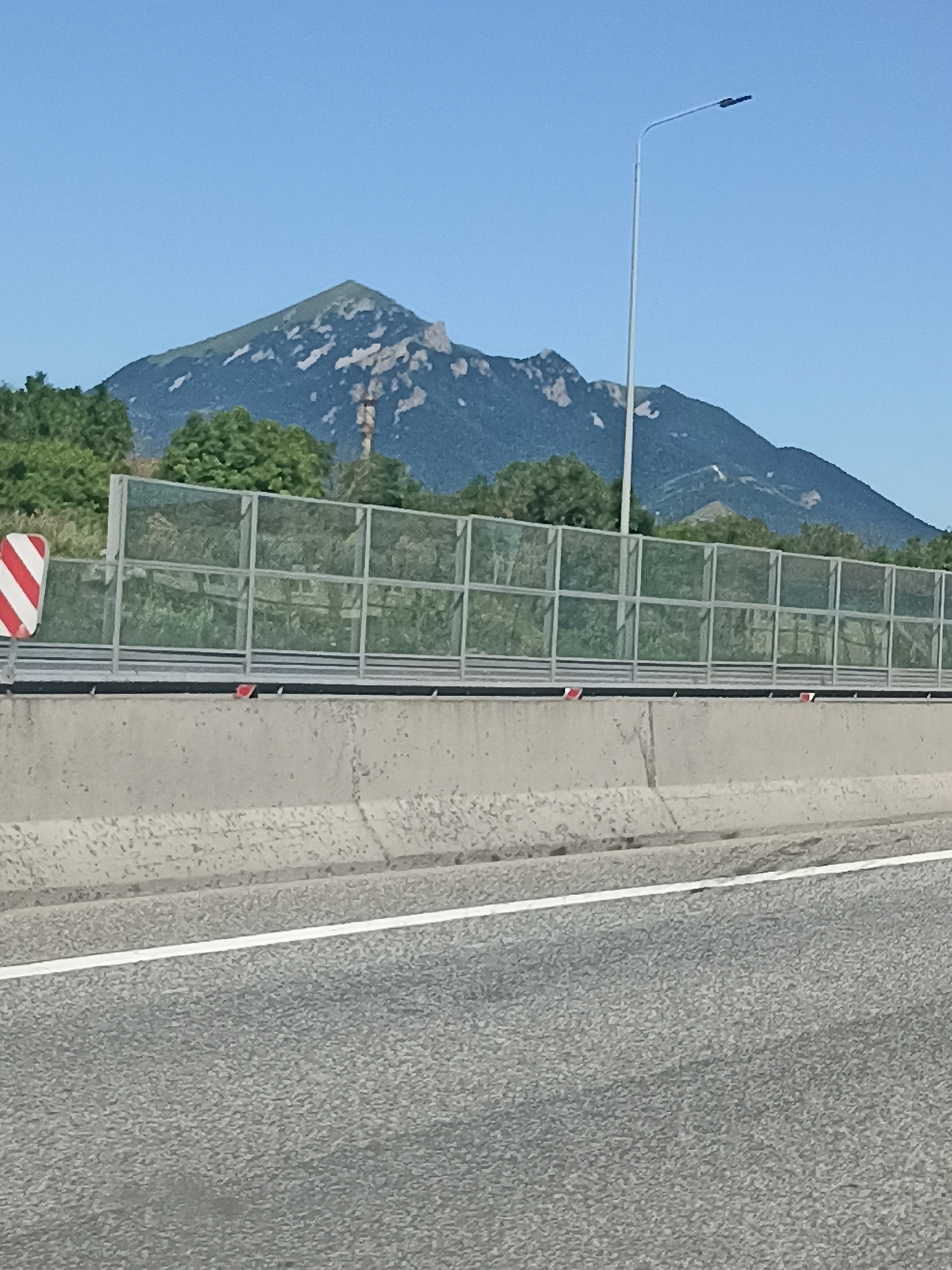 The image captures a scenic view of a mountain range under a clear blue sky, possibly taken from a highway in or near Inozemtsevo, Russia. In the foreground, a section of an asphalt road with a white lane marker is visible. Behind the road, a concrete barrier separates it from a sound barrier composed of metal posts and glass panels. Beyond the sound barrier, lush green trees form a dense line, obscuring the lower slopes of the mountains. The mountains themselves are rugged and imposing, with patches of rocky, exposed terrain contrasting with areas of green vegetation. A tall, modern street lamp with a dark fixture stands prominently in the middle ground, to the right of the mountain peak. The overall impression is one of a natural landscape juxtaposed with man-made infrastructure.