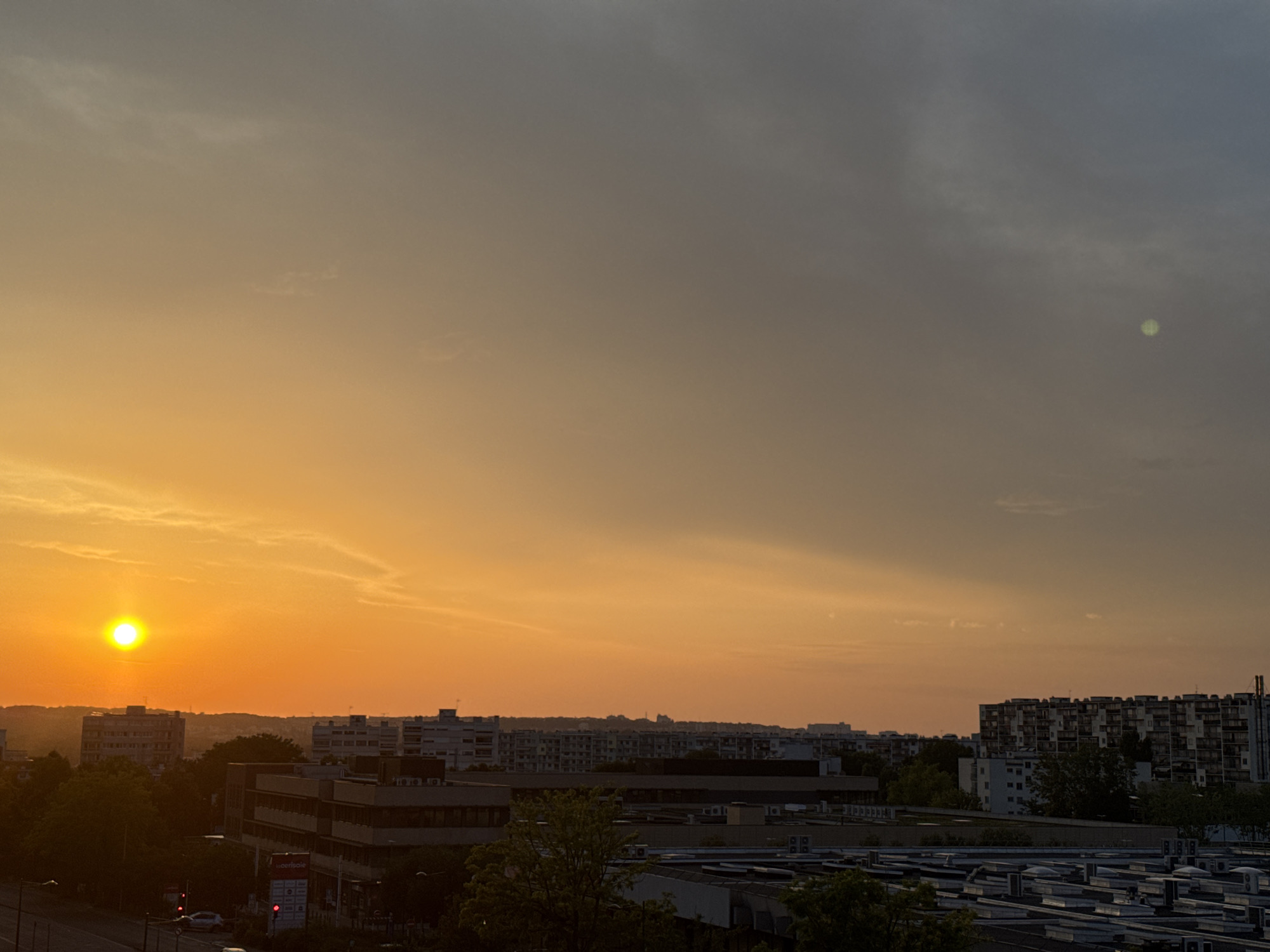 A wide shot captures a city skyline during a sunset in an unknown location. The sky is a gradient of deep orange and gray, with a bright orange sun prominently displayed on the left side. A soft glow emanates from the sun, casting a warm light over the scene. In the foreground, buildings and trees are silhouetted against the vibrant sky. A street with cars and traffic lights is visible in the lower left portion of the image. The overall impression is one of a serene urban sunset.